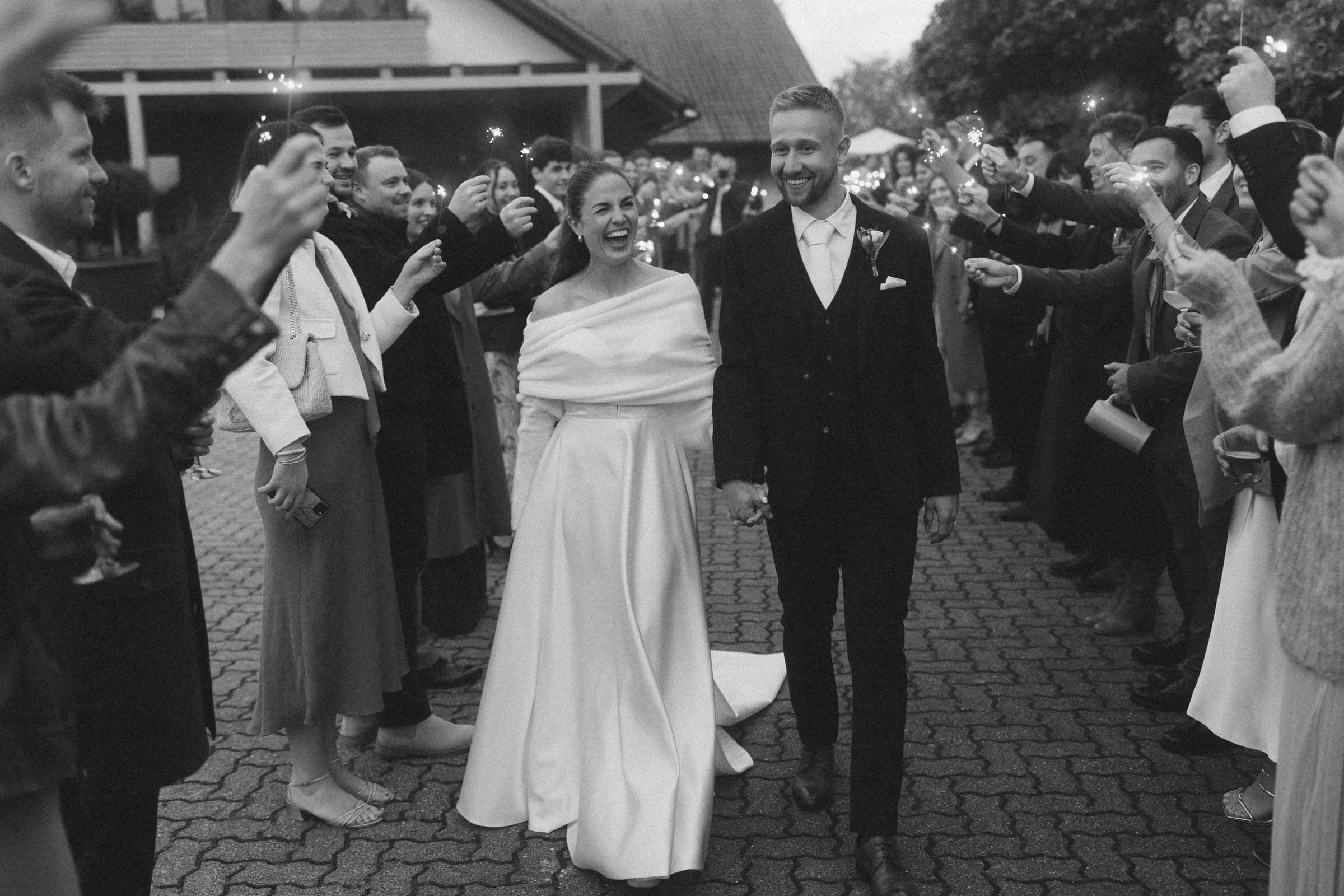 Braut und Bräutigam gehen Hand in Hand durch einen gestreiften Gassenabschnitt, umgeben von Feiernden mit Feuerwerksfunken in der Nacht, bei einer Hochzeit.