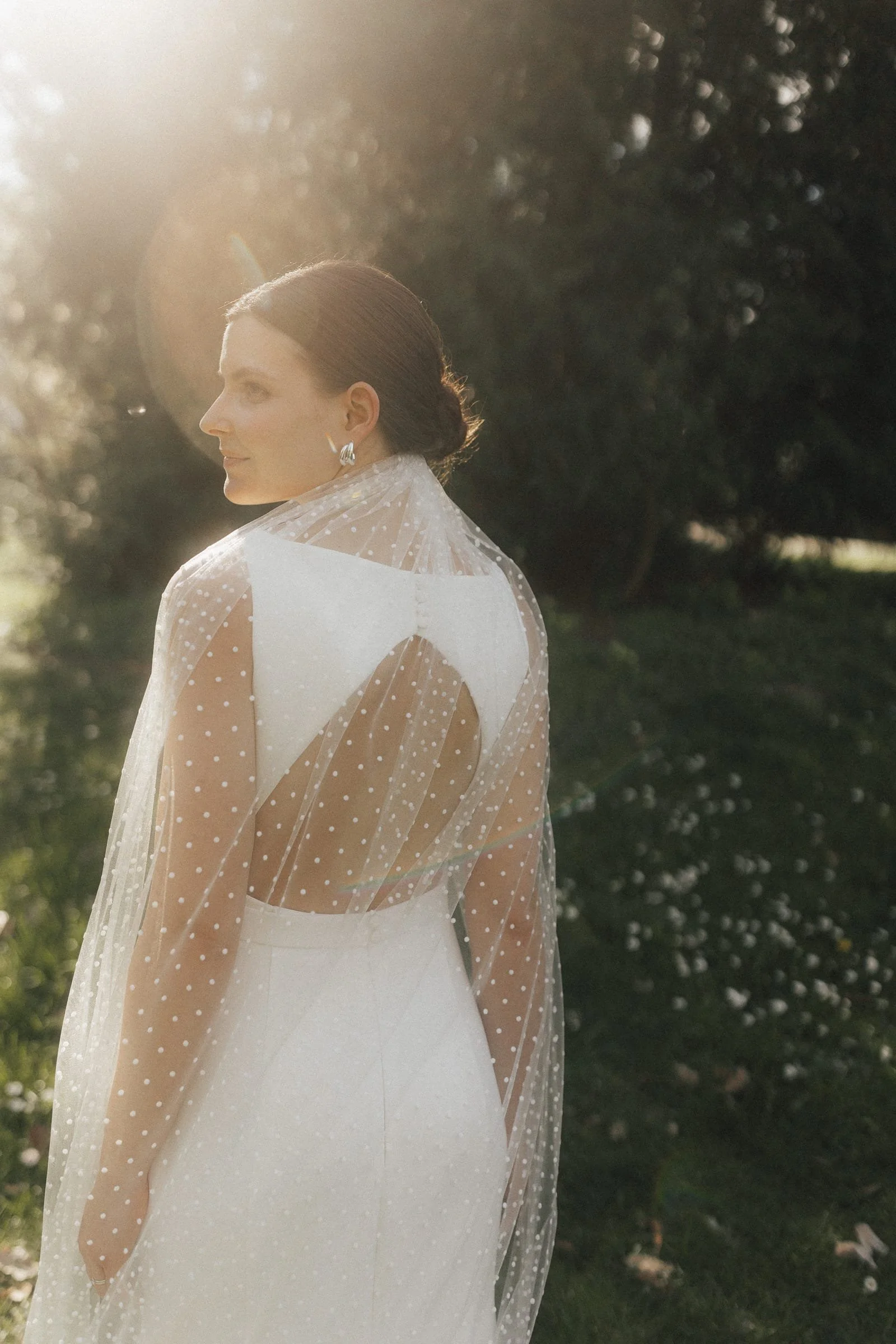 Eine Frau in einem eleganten weißen Kleid mit einer transparenten, gepunkteten Schleife, die auf einer Rasenfläche bei Sonnenuntergang steht, umgeben von Bäumen.