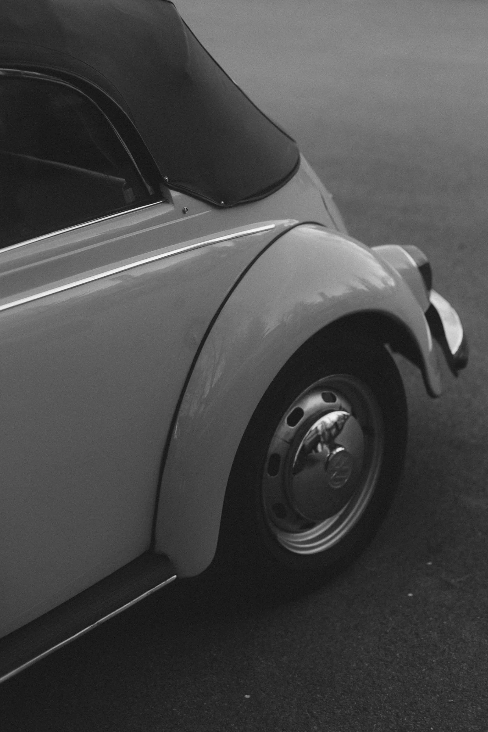 Nahaufnahme eines klassischen Autos mit silberner Farbe, schwarzem Dach und silbernem Rad mit VW-Logo, auf dunklen Asphaltfläche.