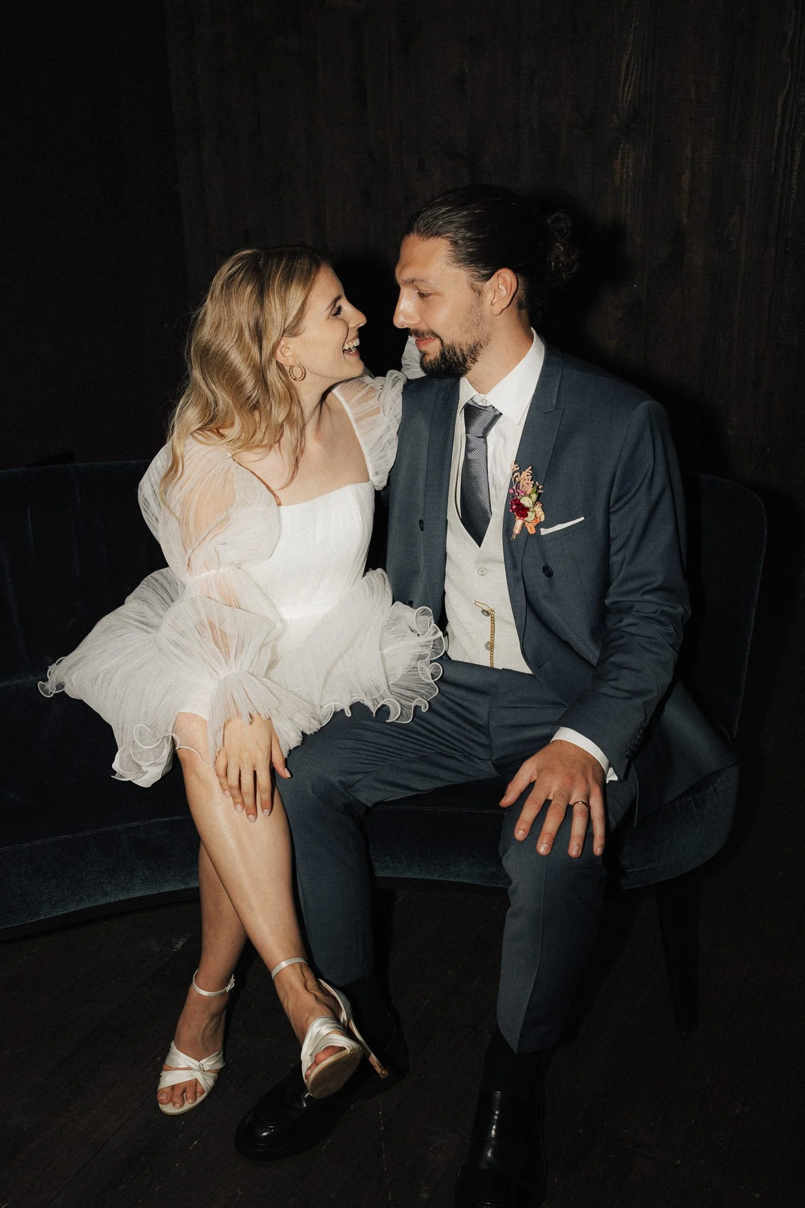 Ein Paar sitzt auf einem schwarzen Sofa, sieht sich liebevoll an, trägt formelle Kleidung, vermutlich bei einer Hochzeit oder ähnlichen Feier.