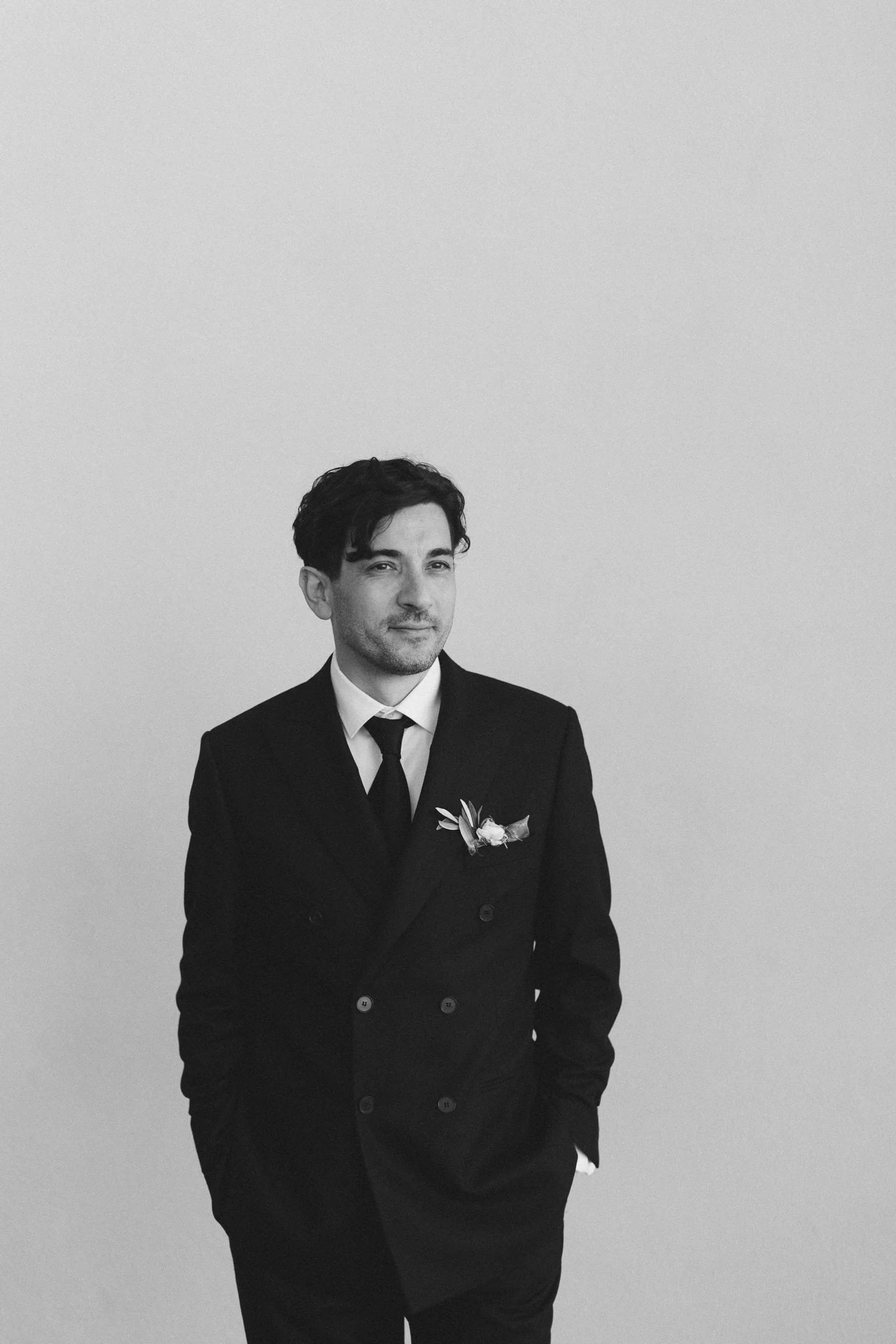 Schwarz-weiß-Foto eines jungen Mannes in formeller dunkler Kleidung, mit Anzug und Krawatte, vor einem schlichten, hellgrauen Hintergrund. Er trägt eine kleine Blume an der Jacke. Er hat dunkle, lockige Haare und einen leichten Bart.