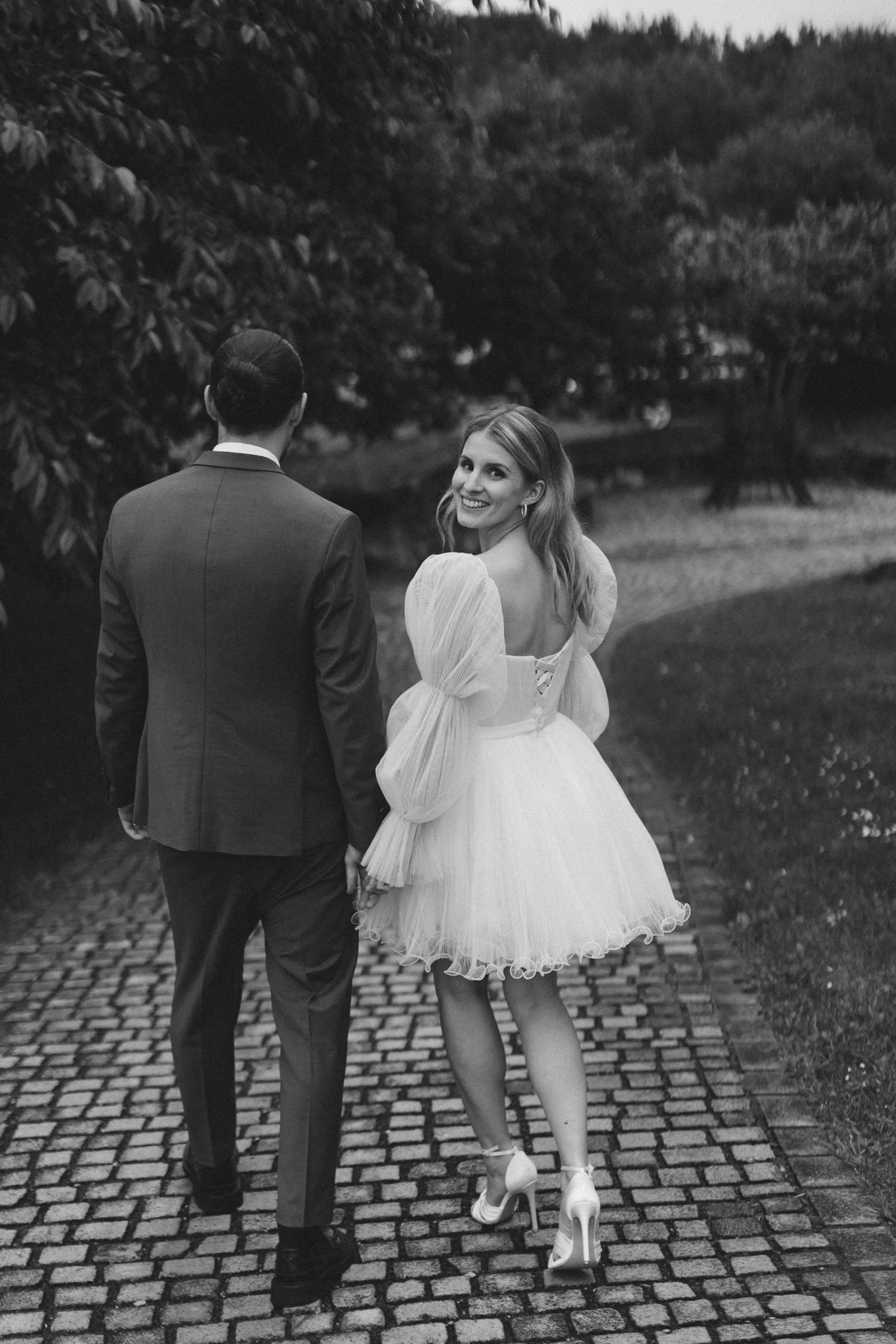 Ein glückliches Paar, ein Mann in einem Anzug und eine Frau in einem eleganten Brautkleid, geht Hand in Hand auf einem Kopfsteinpflasterweg in einem Park.