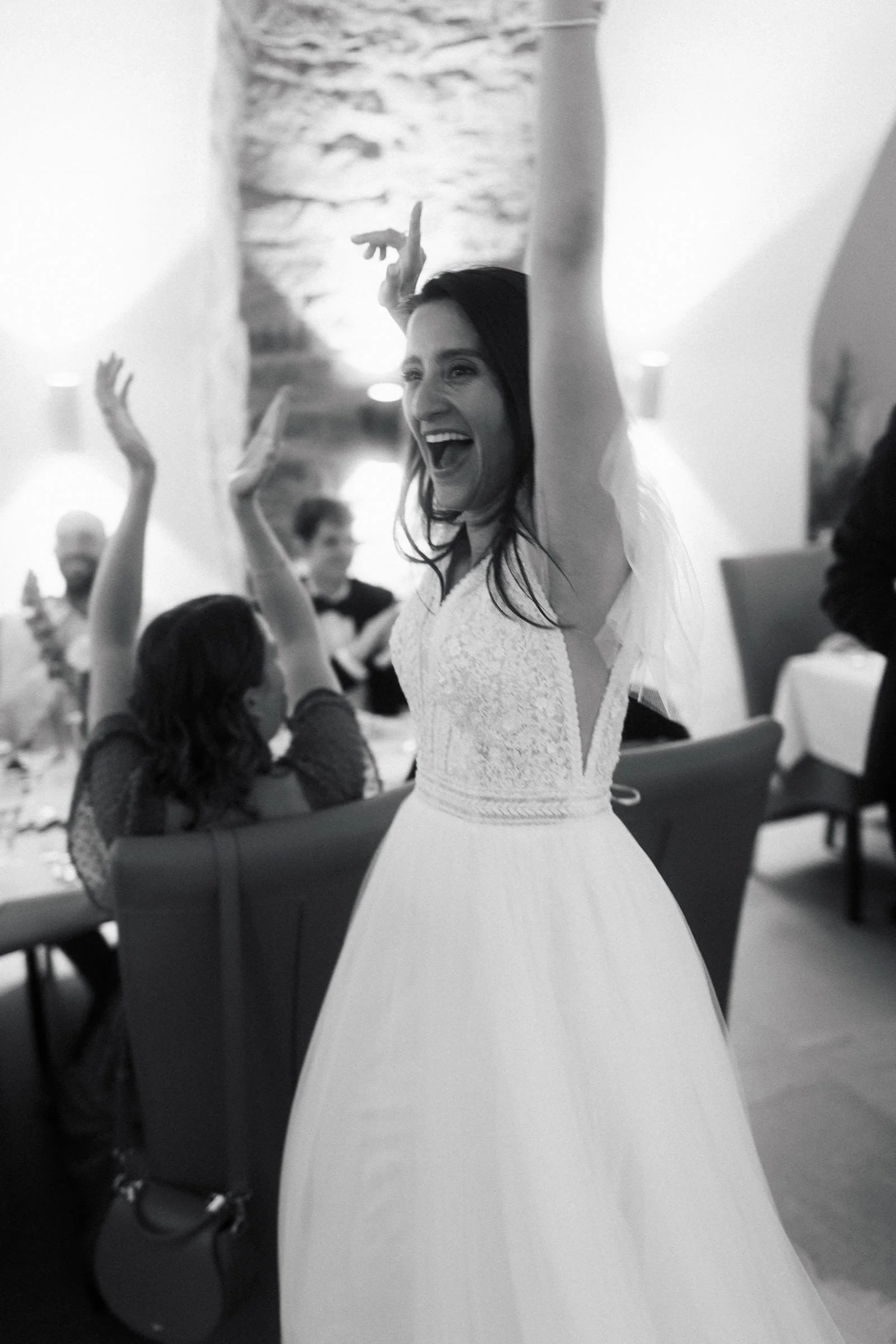 Jubilarin in einem Brautkleid feiert laut lachend während einer Feier, andere Gäste im Hintergrund mit Armen in die Luft.