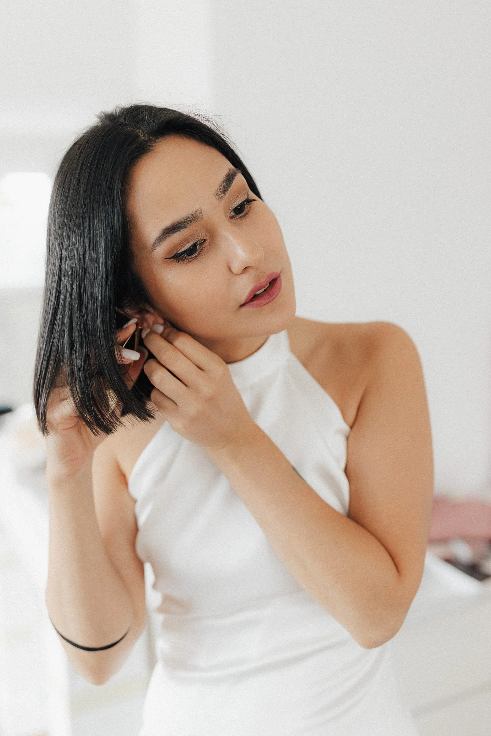 Eine Frau mit dunklen Haaren, die sich Schmuck in das Ohr steckt, trägt ein weißes elegantes Kleid und schaut nach unten.