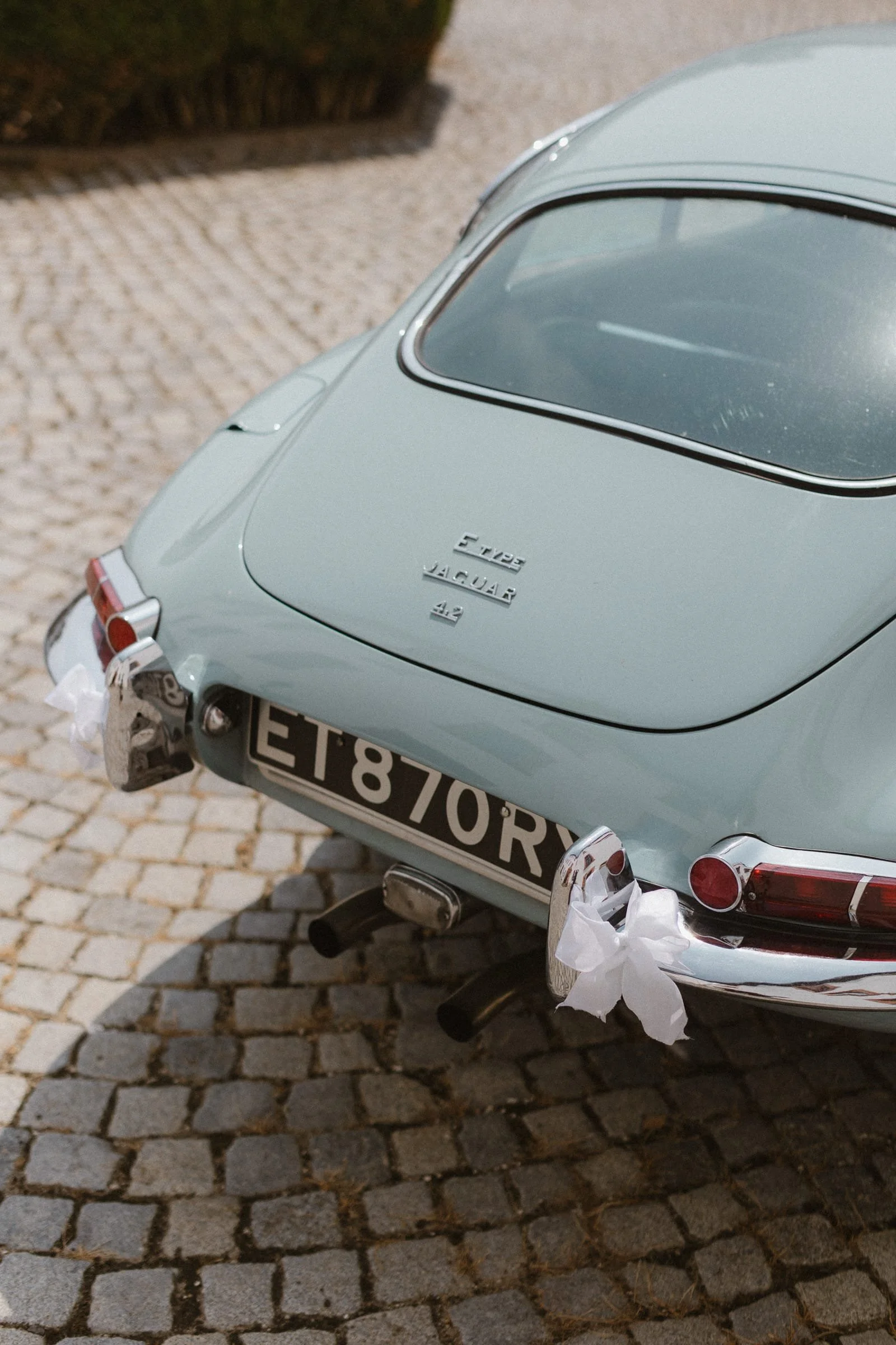 Nahaufnahme eines klassischen Jaguar E-Type Autos, das auf einer Kopfsteinpflasterstraße geparkt ist, mit weißen Blumen an den Stoßstangen.