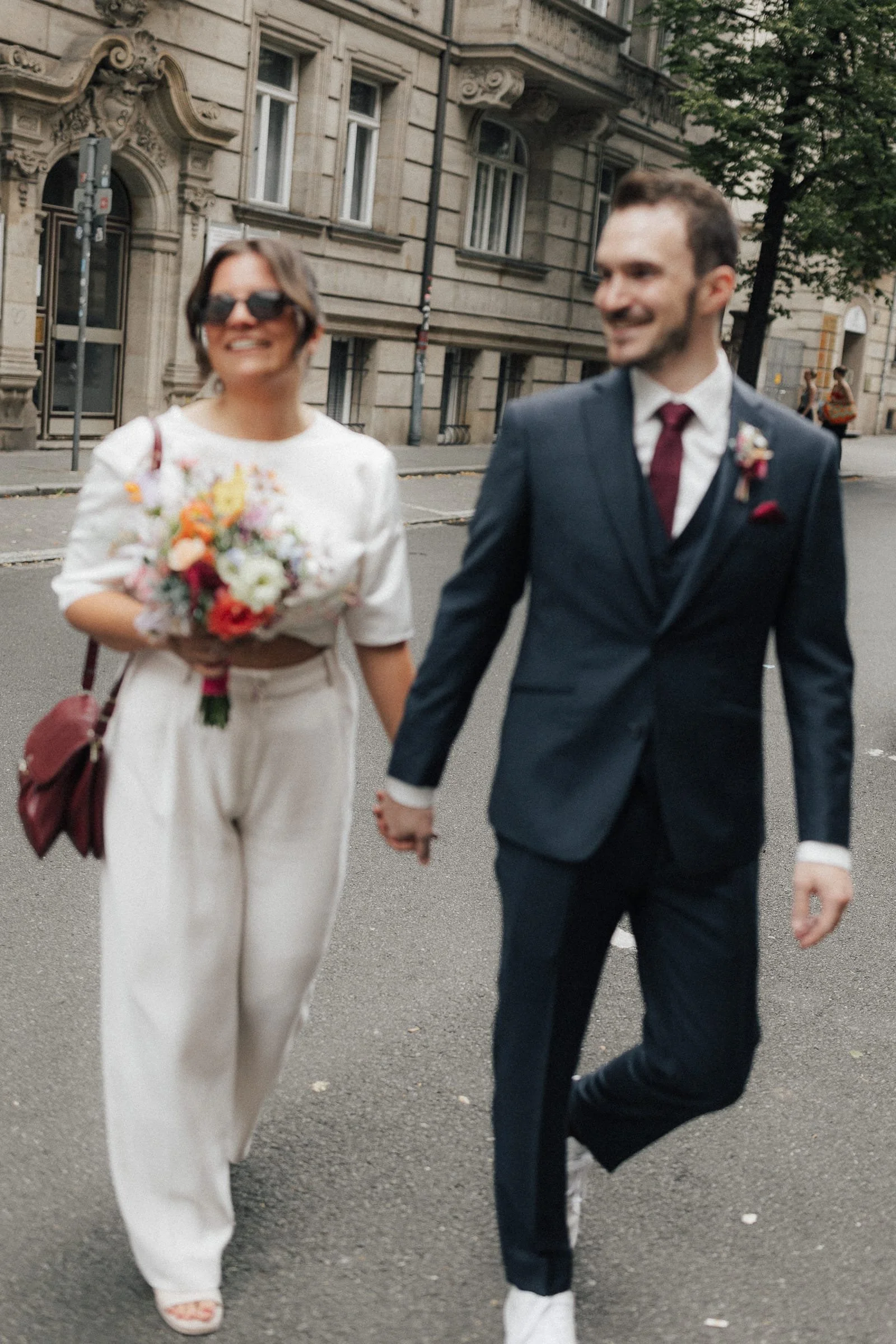 Ein Paar, vermutlich Braut und Bräutigam, hält Händchen beim Spaziergang auf der Straße in einer Stadt, mit historischen Gebäuden im Hintergrund.