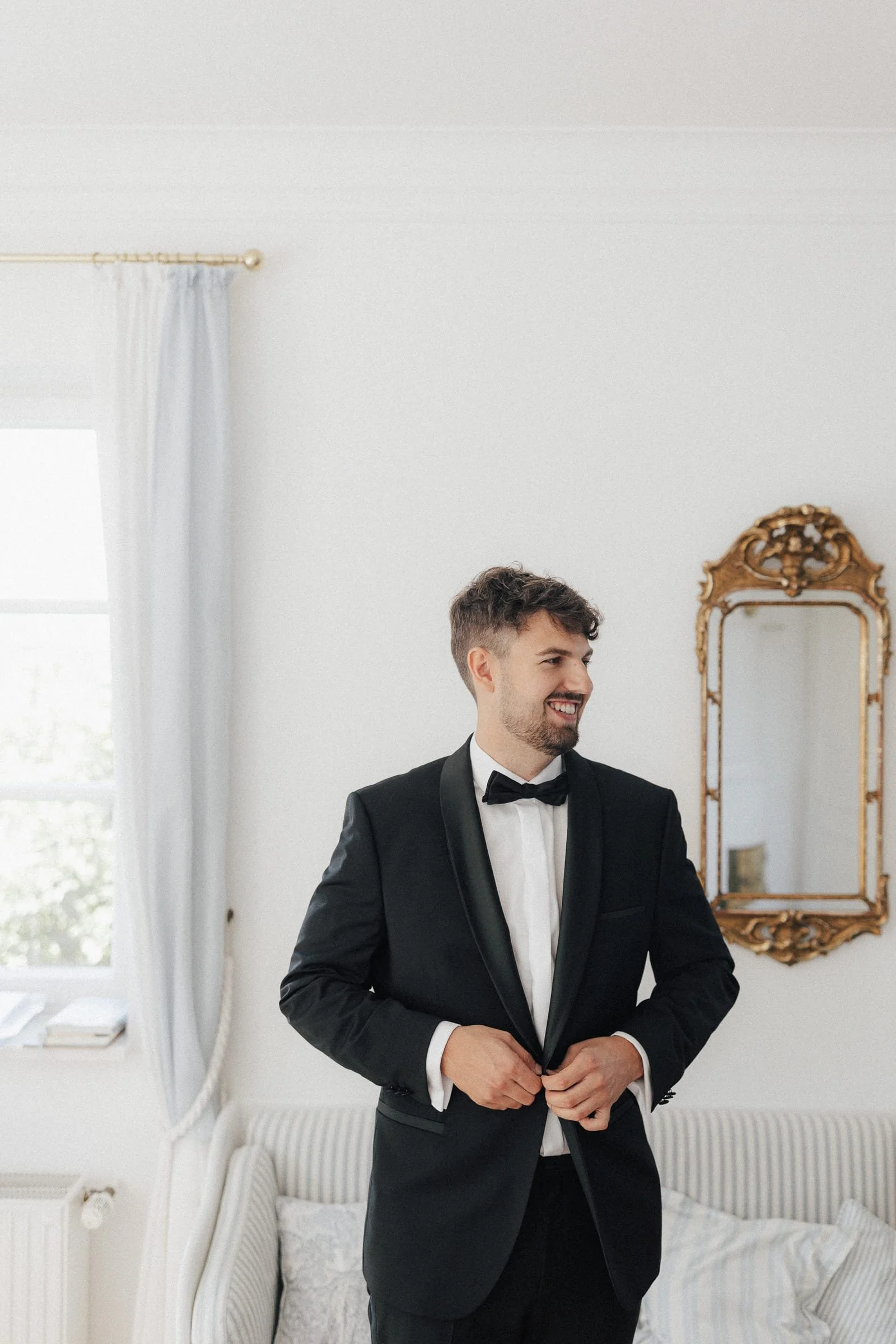 Ein Mann in einem schwarzen Anzug und Fliege, der sich vor einem Spiegel anzieht, steht in einem hellen Raum mit weißen Wänden und einem Sofa.