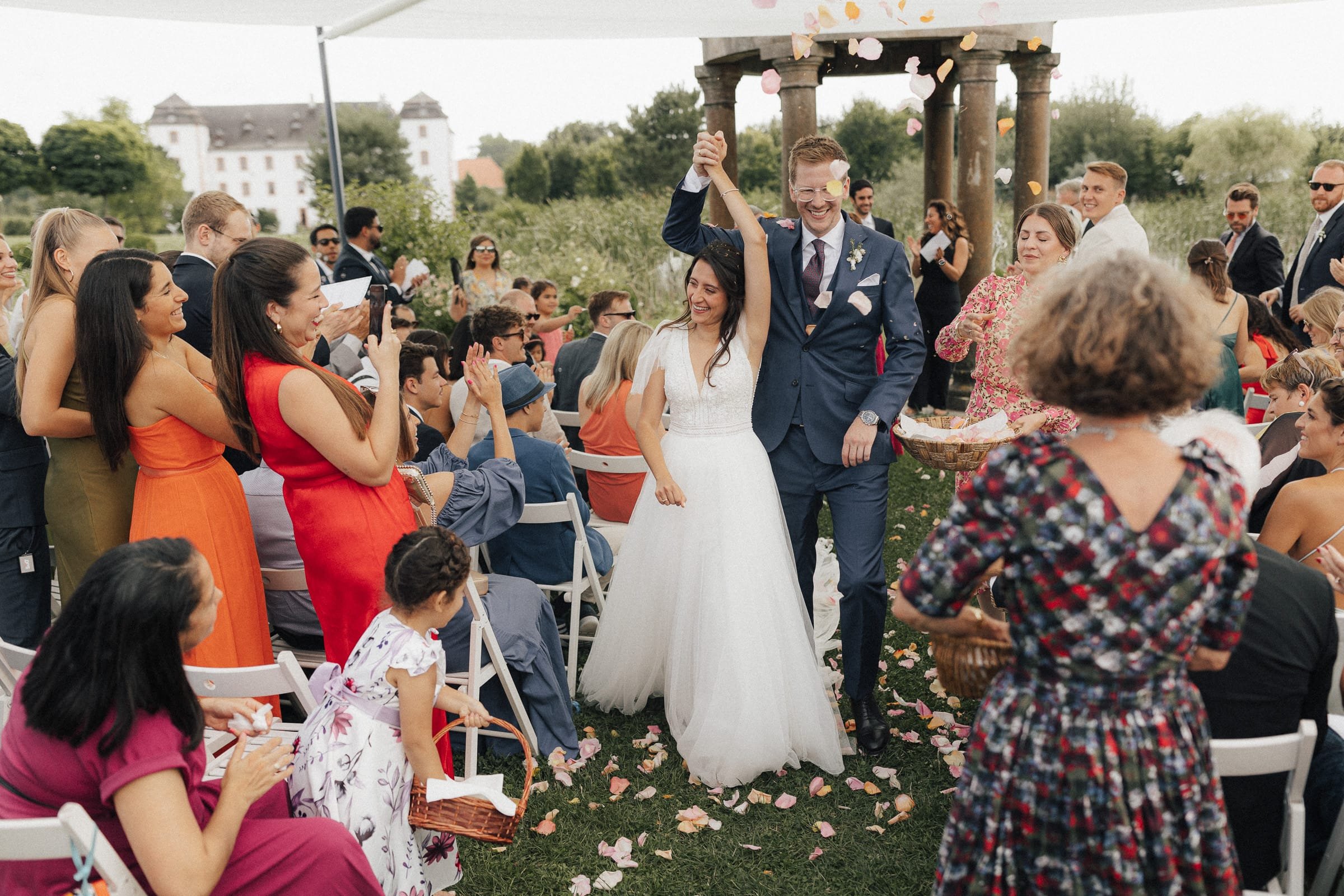 Braut und Bräutigam laufen Hand in Hand durch eine Feiergesellschaft, während Konfetti geworfen wird, im Freien bei einer Hochzeit.