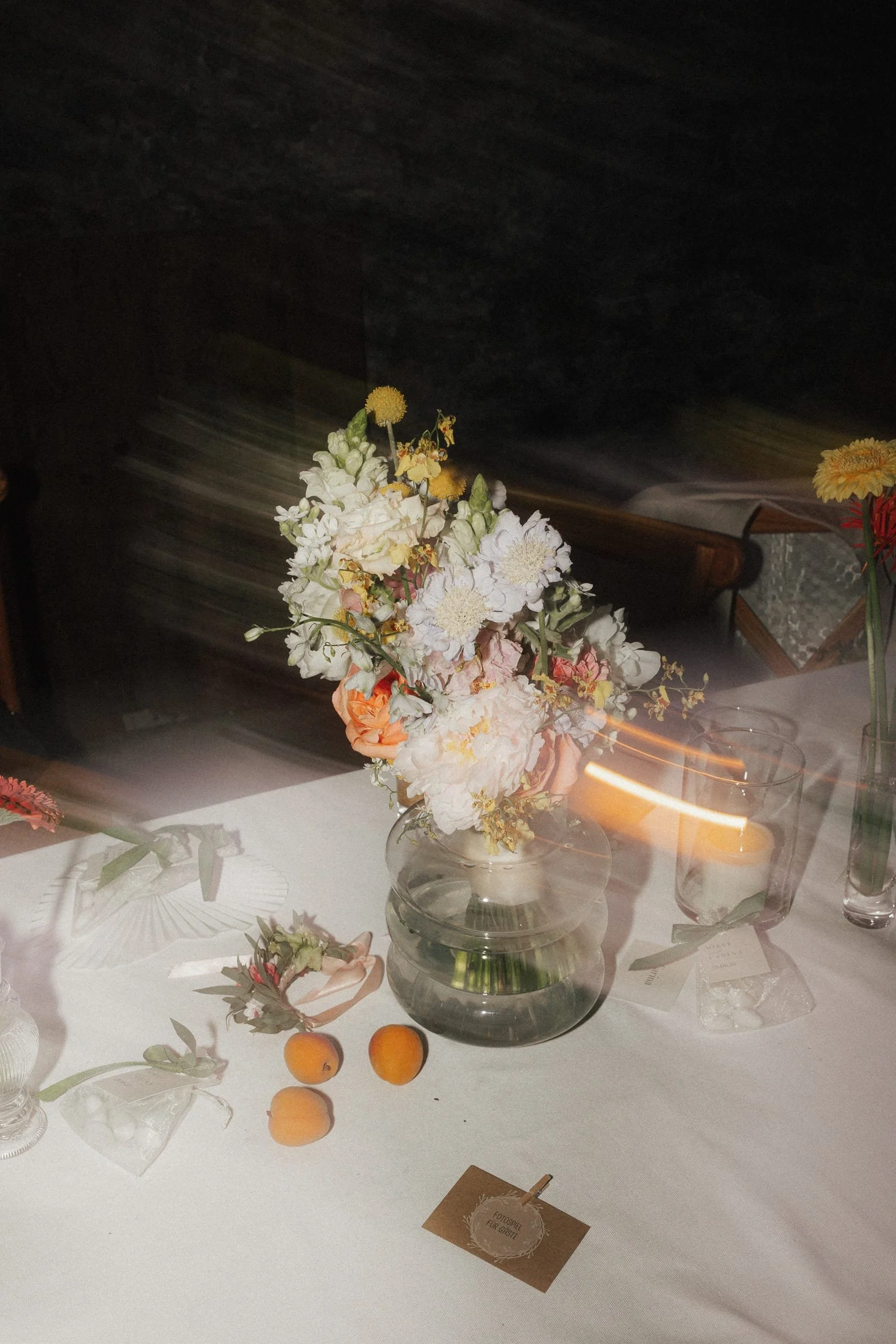 Blumenvase mit weißen und pastellfarbenen Blumen auf einem weißen Tisch, umgeben von Kerzen, Früchten und Dekorationsgegenständen, bei einer Veranstaltung oder Feier.