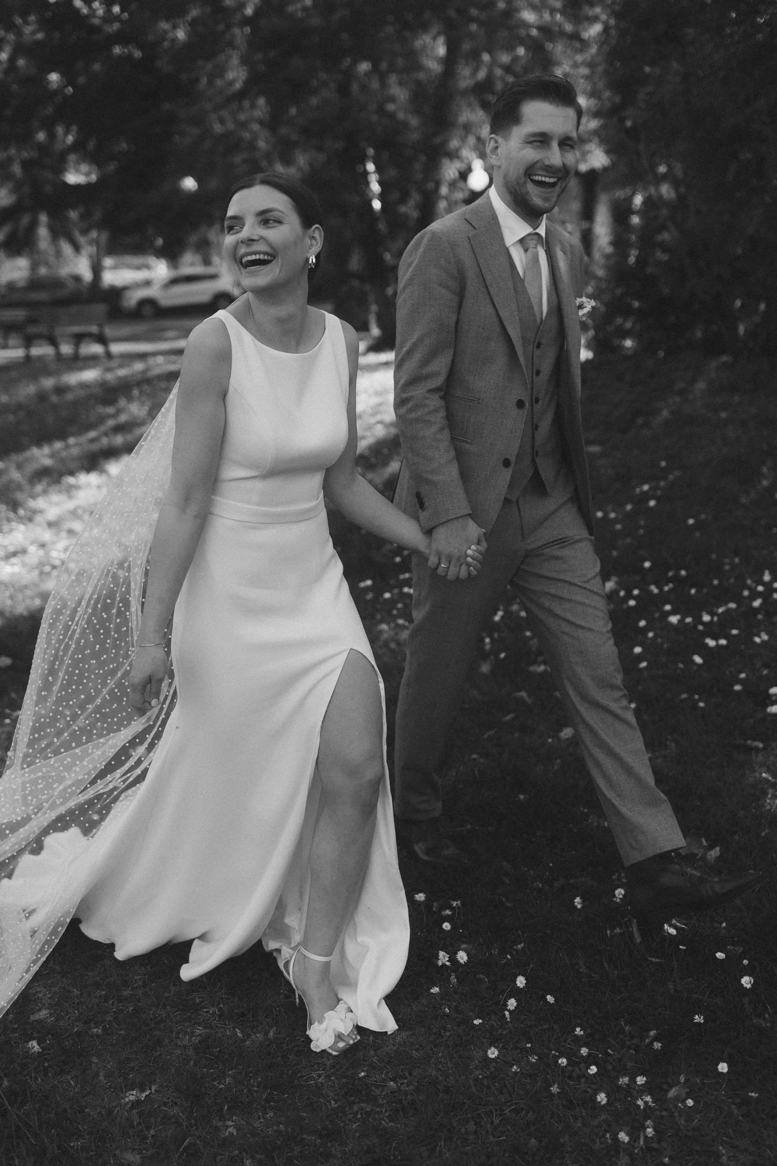 Ein glückliches Brautpaar in formeller Kleidung spaziert im Park, hält Händchen, umgeben von Bäumen und Blumen, schwarz-weiß fotografiert.