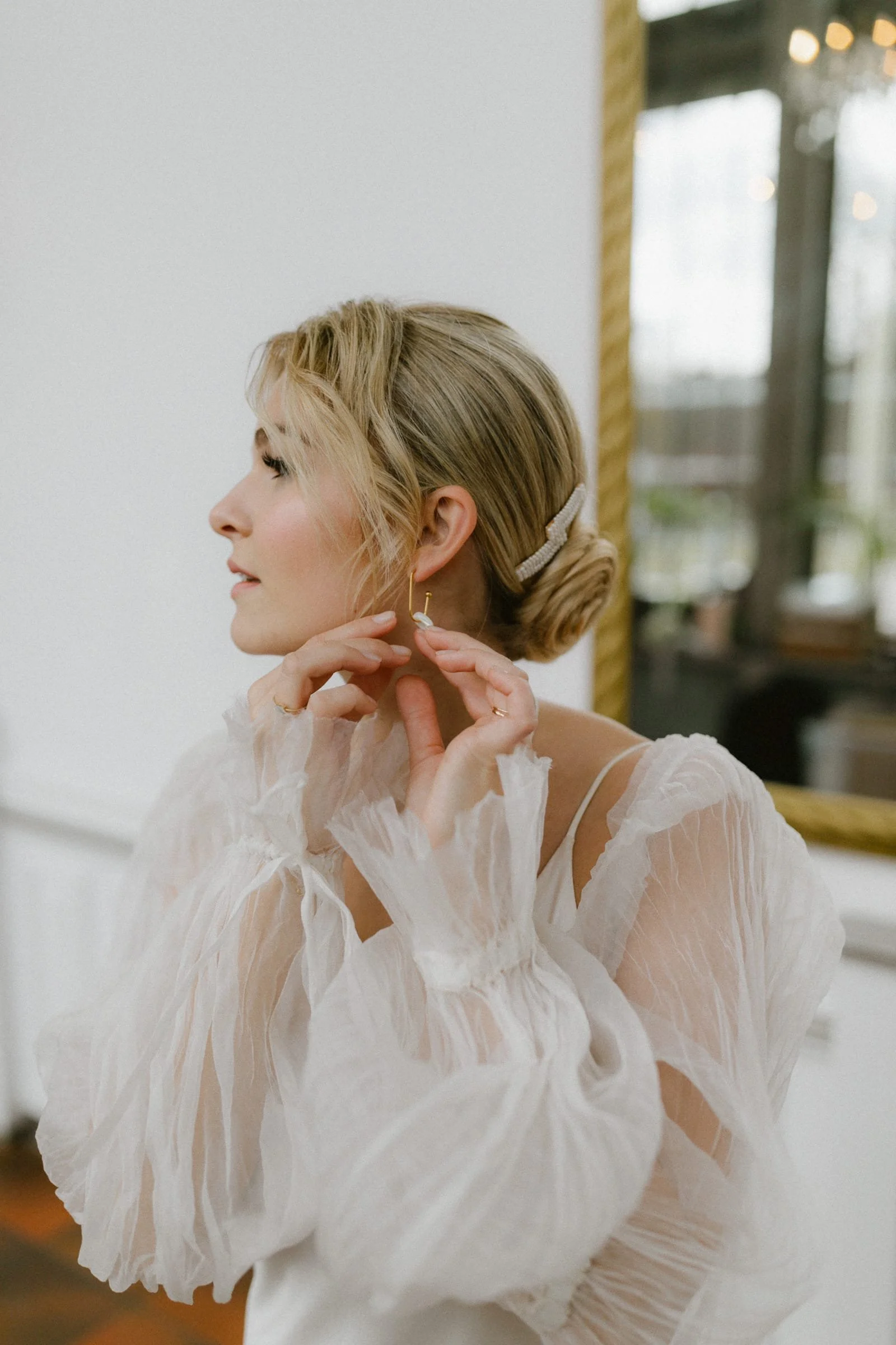 Eine Frau mit blonden Haaren in einem weißen, transparenten Kleid, die Ohrringe anlegt, steht vor einem Spiegel.