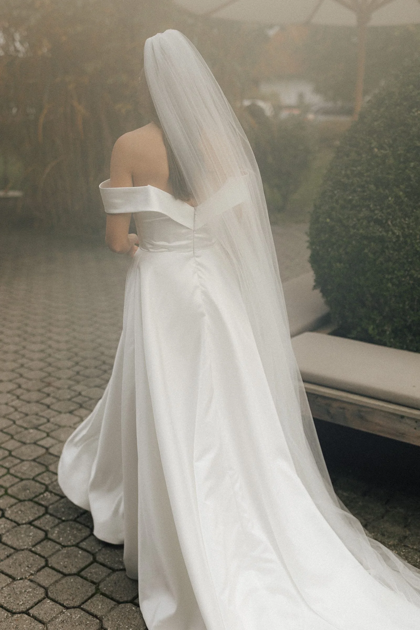 Braut in langem weißen Hochzeitskleid mit Schleier, von hinten fotografiert, steht auf Pflastersteinweg neben einer Bank, im Hintergrund Büsche und Gebäude.