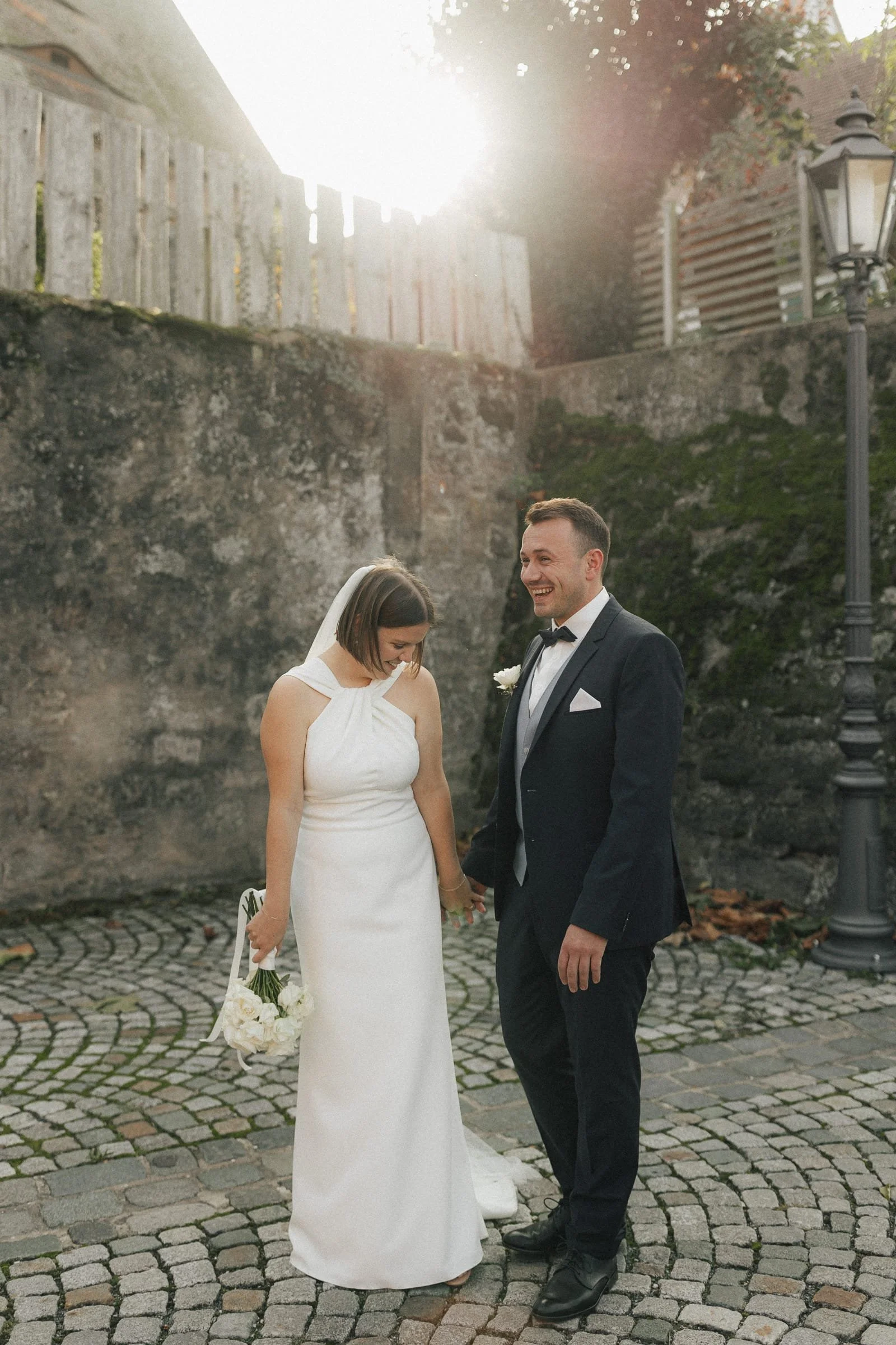 Ein Brautpaar bei einer Hochzeit, die Frau in einem weißen Kleid, der Mann in einem schwarzen Anzug, hält Händchen und lacht. Die Szene findet im Freien auf einem gepflasterten Weg statt. Es ist sonnig mit Lichtstrahlen im Hintergrund.