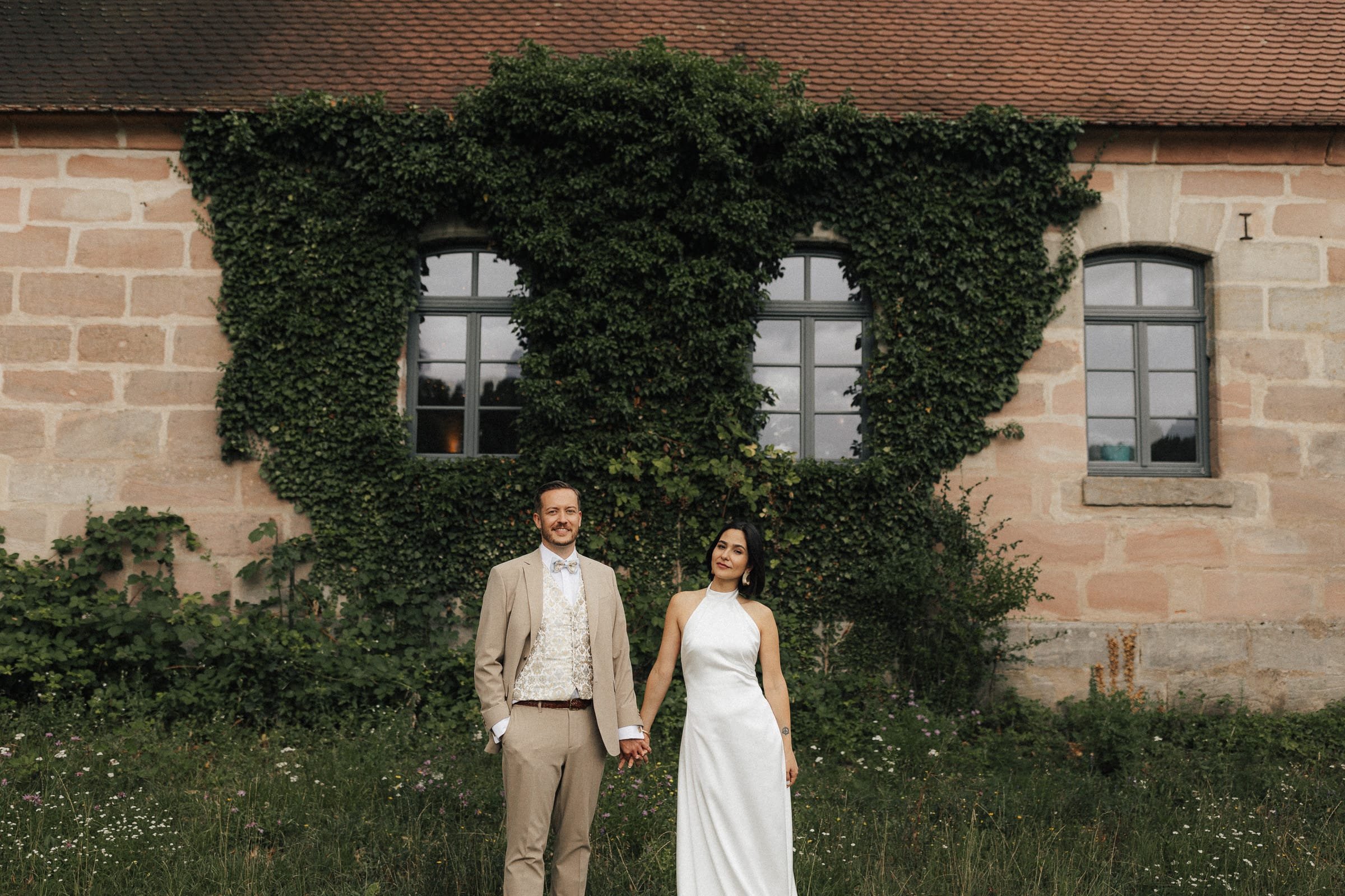 Ein verliebtes Paar in Hochzeitskleidung hält sich an einem Tag im Grünen vor einem historischen Backstein-Gebäude mit Efeu bewachsen auf.