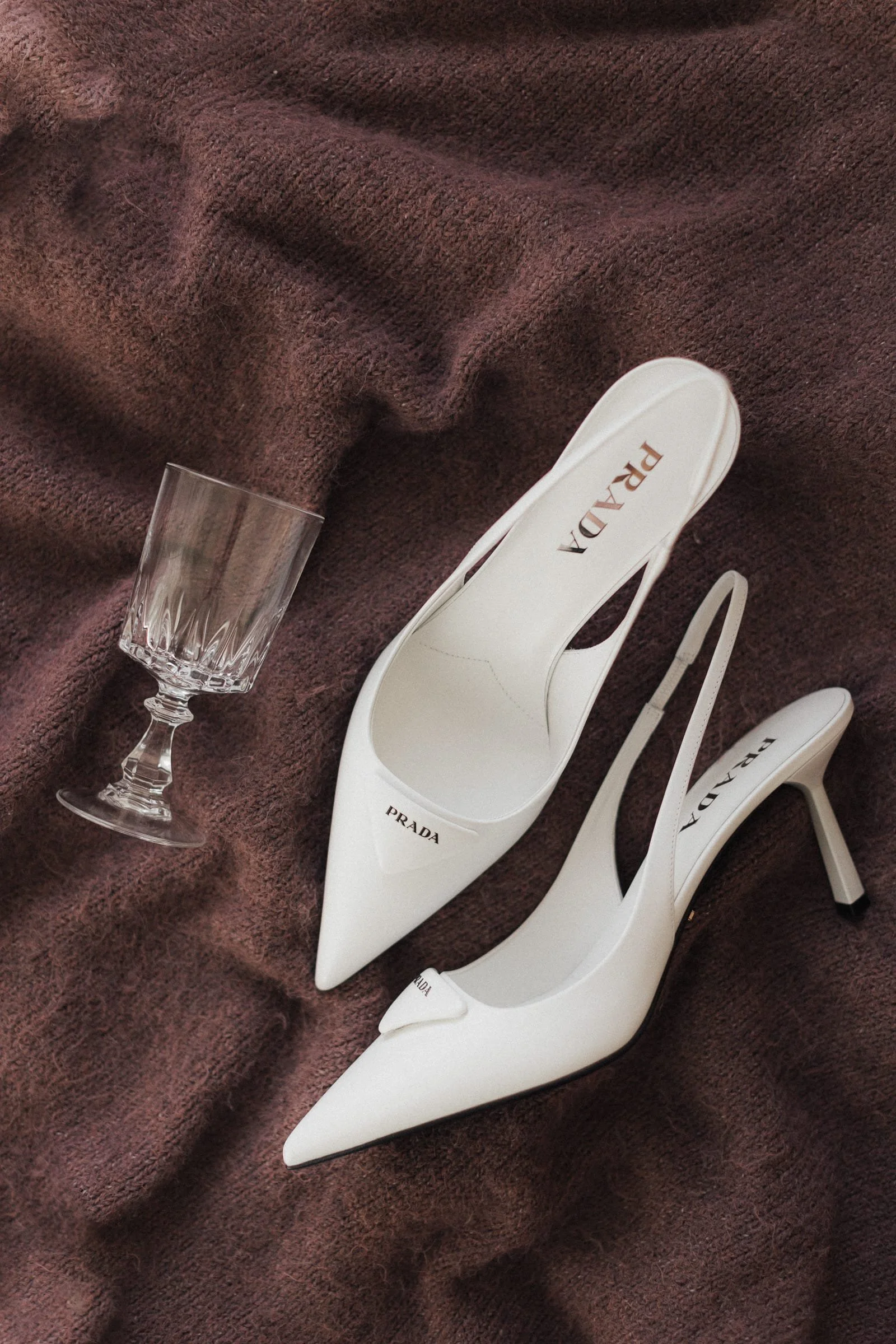 Weißes Paar High Heels von Prada auf burgunderfarbenem Stoff, neben einer leeren Kristall-Glas