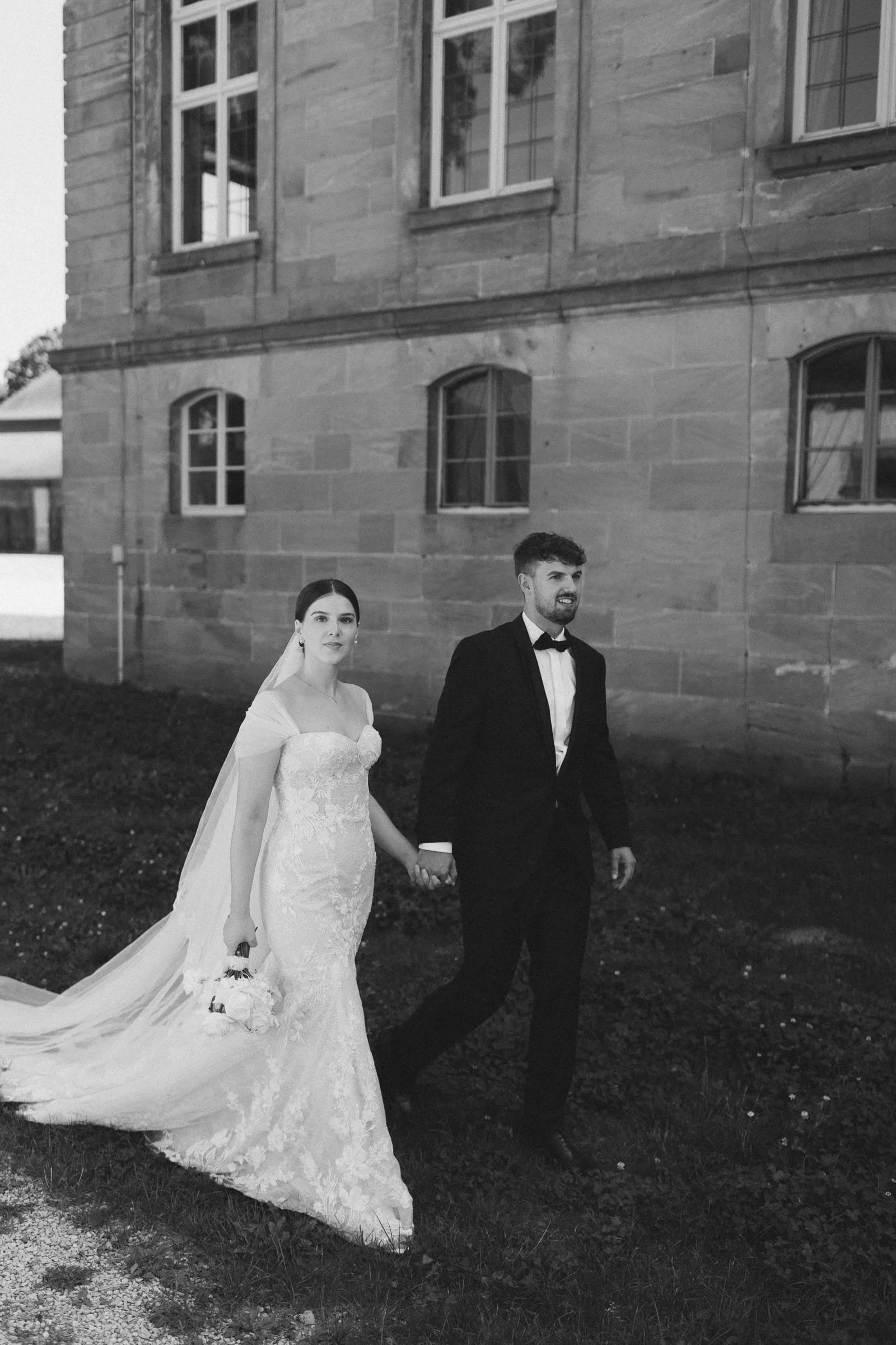 Ein Brautpaar in formeller Kleidung, die Hand in Hand vor einem alten Stein-Gebäude  spazieren gehen.