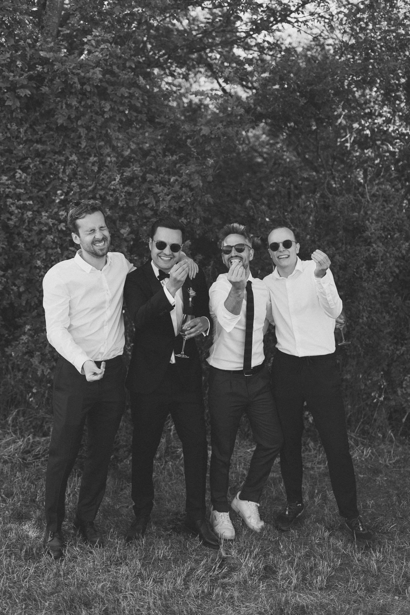 Fünf Männer in formeller Kleidung lachen und feiern im Freien, einige mit Sonnenbrillen, auf einer Wiese vor Bäumen, in Schwarz-Weiß aufgenommen.
