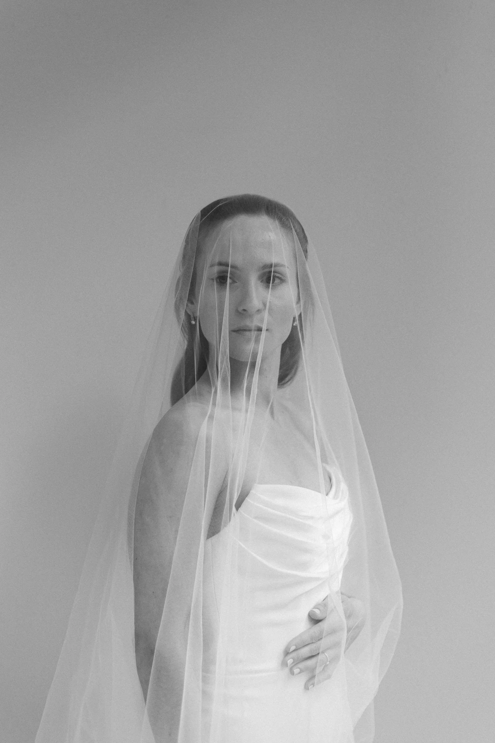 Schwarz-weiß-Bild einer eleganten Braut im Brautkleid mit Schleier vor einem neutralen Hintergrund.