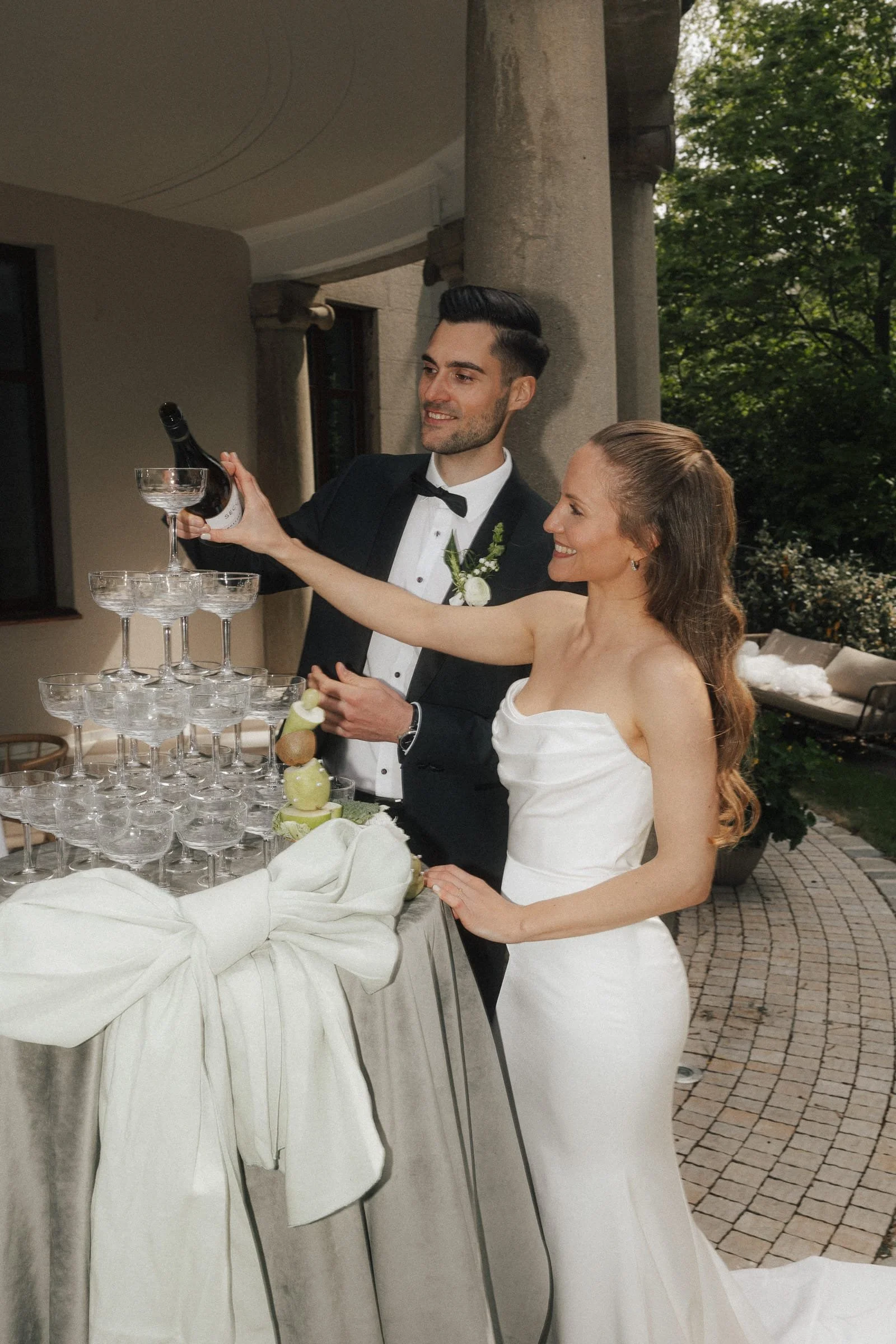 Ein Brautpaar beim Anstoßen bei einer Hochzeit, sie trinken Champagner aus einem hohen Weinglas, der Tisch ist mit einem weißen Tuch bedeckt und es sind leere Gläser gestapelt.