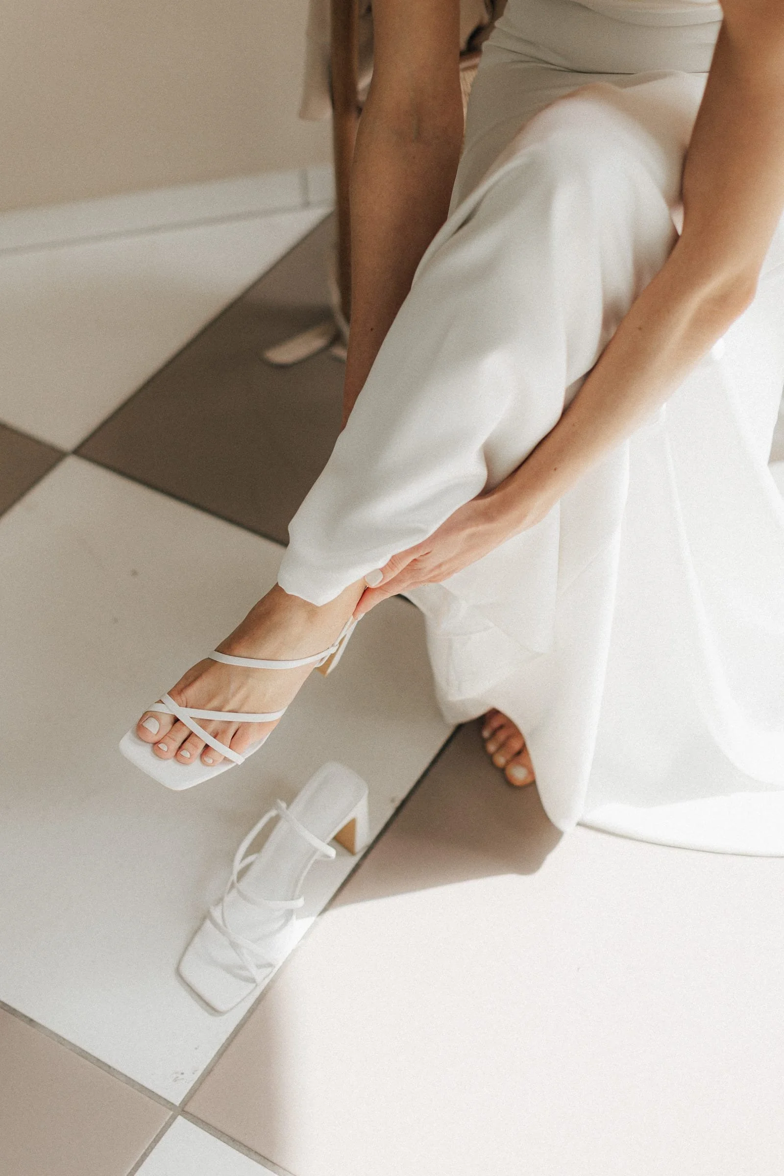 Eine Braut zieht weiße High Heels an, die auf einem Fliesenboden stehen, während sie im Spiegel betrachtet wird.