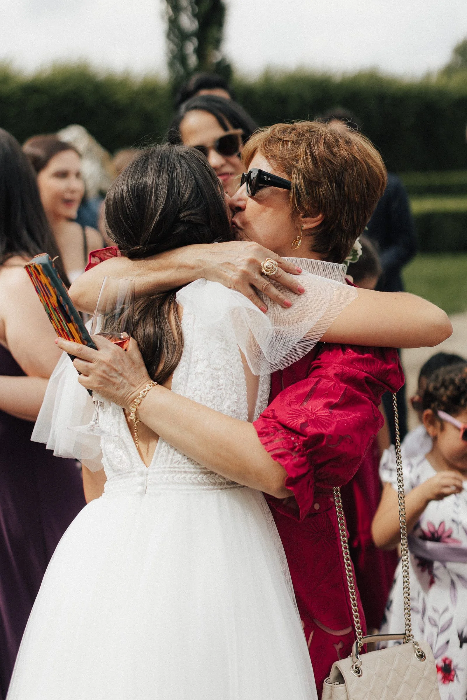 Zwei Frauen umarmen sich herzlich bei einer Feier im Freien, eine trägt ein Hochzeitskleid, die andere trägt ein rotes Kleid.