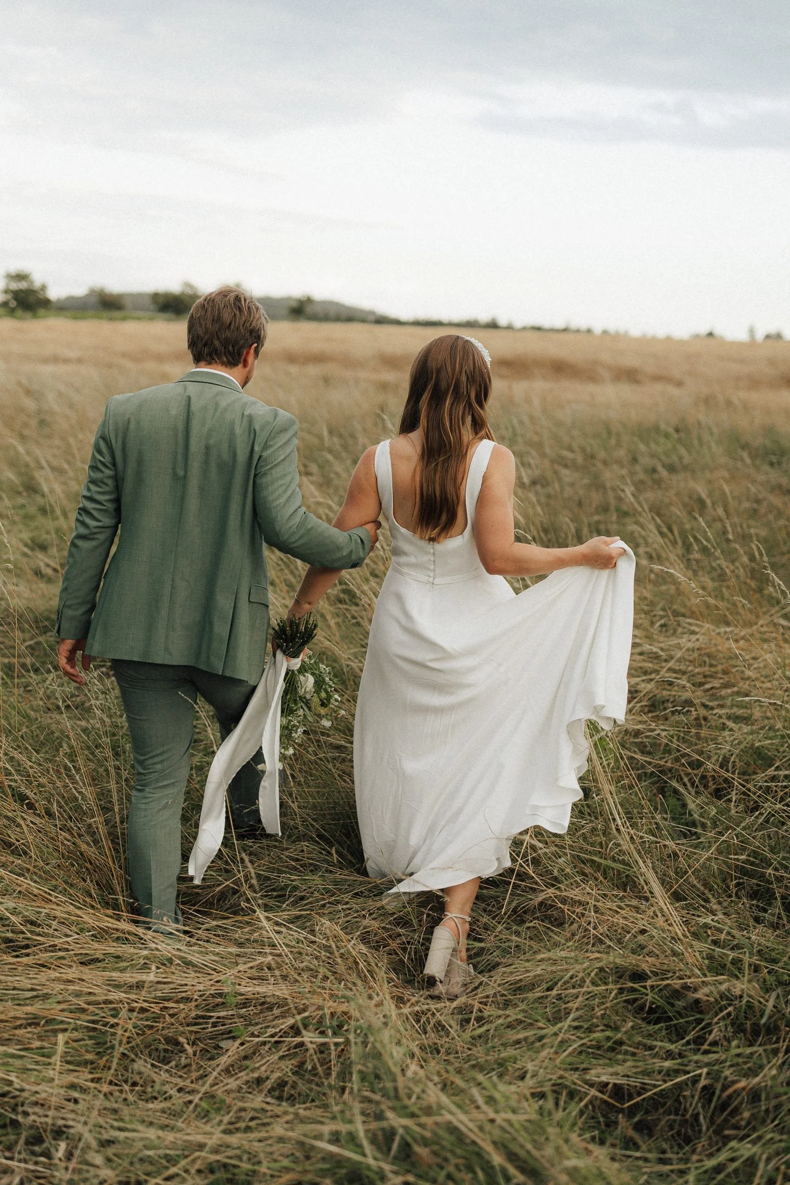 Ein Paar läuft gemeinsam durch ein Feld, das Braut und Bräutigam bei einer Hochzeit. Die Frau trägt ein weißes Hochzeitskleid und hohe Schuhe, während der Mann einen Anzug trägt.