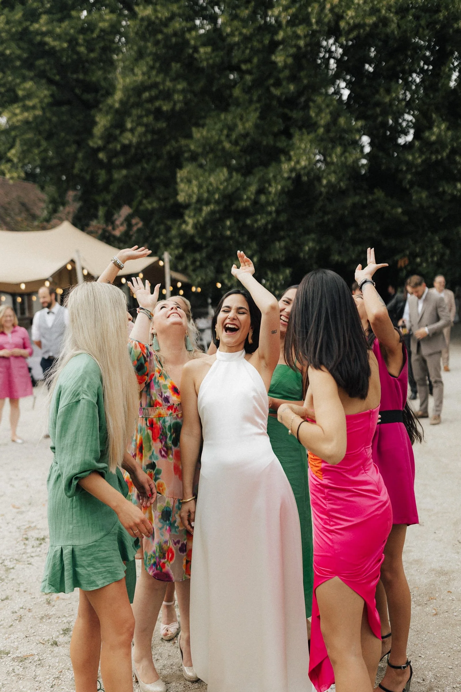 Gruppe lachender Frauen bei einer Outdoor-Feier, umgeben von Bäumen und Zelten.