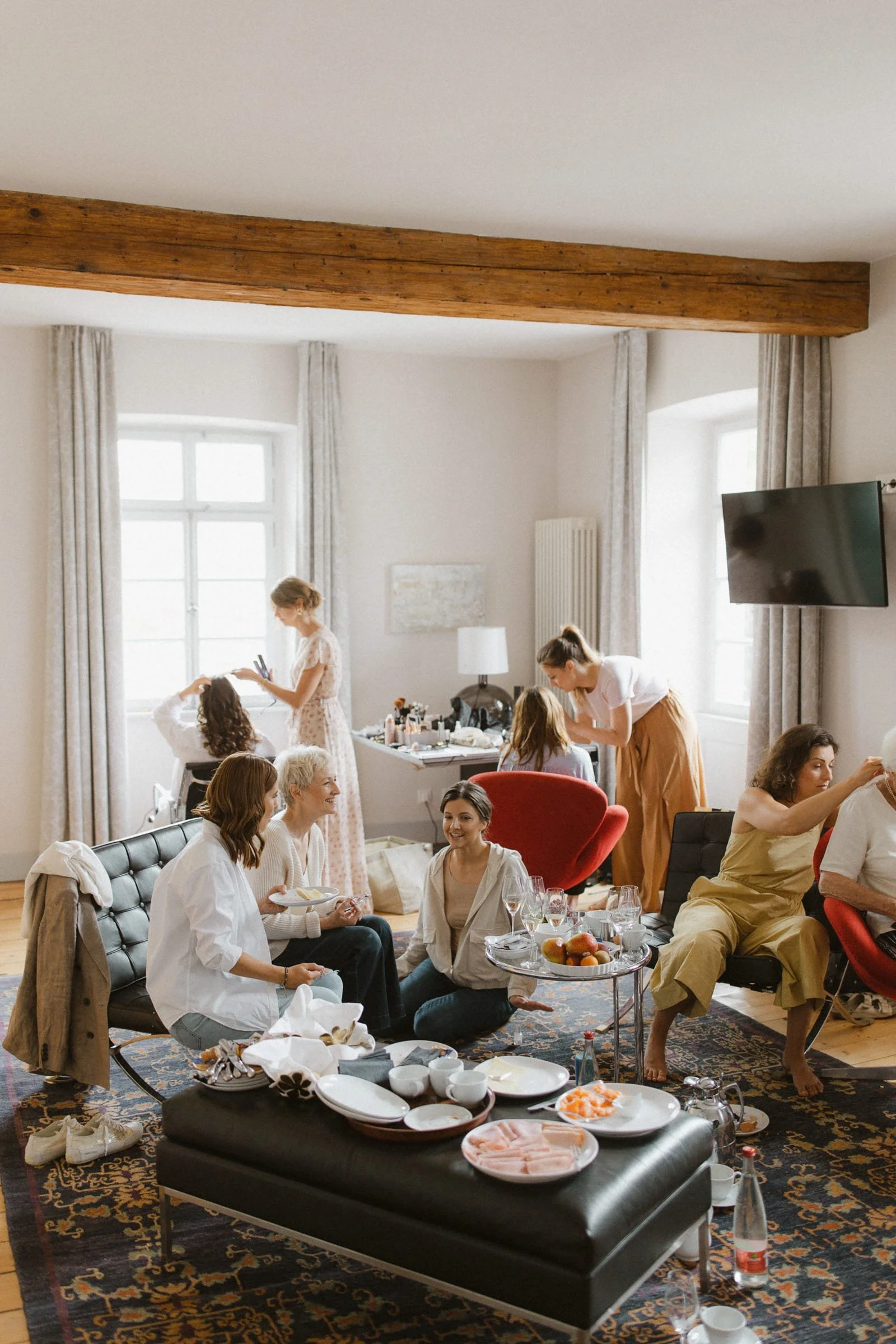 Mehrere Frauen bereiten sich in einem hellen, gemütlichen Raum vor, vermutlich für eine Veranstaltung oder Feier. Sie sitzen, sprechen, schminken und waschen ihre Haare. Es gibt Tische mit Essen und Getränken, und der Raum ist mit großen Fenstern und