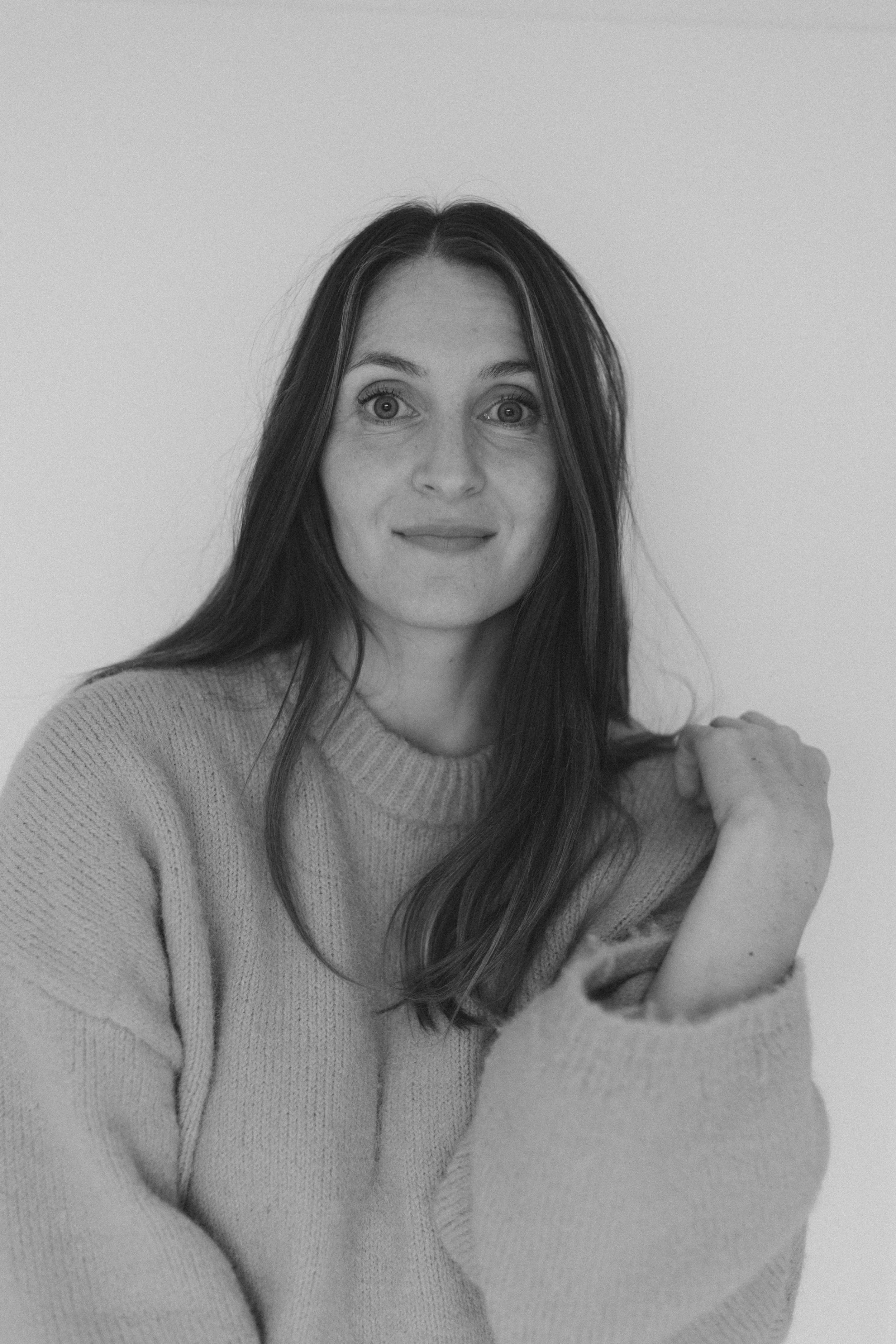 Schwarz-weiß-Foto einer Frau mit langen dunklen Haaren, die ein kuscheliges Pullover trägt und die Hand auf die Schulter gelegt hat.