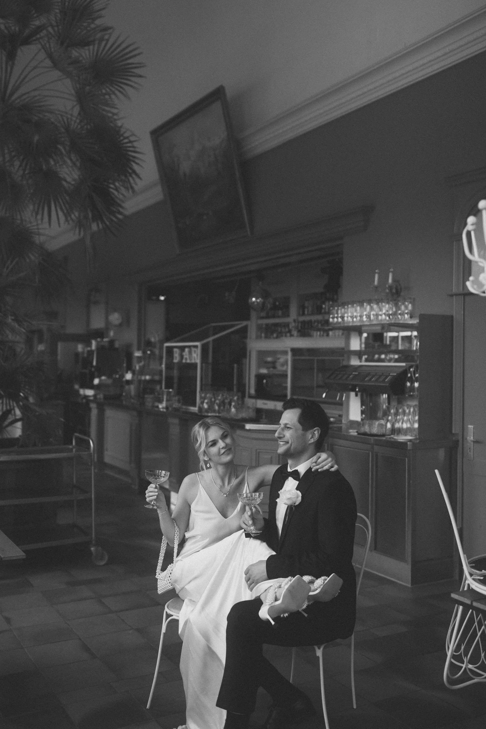 Ein glückliches Paar in formeller Kleidung, vermutlich während einer Hochzeitsfeier, sitzt in einer Bar mit einem Drink in der Hand. Die Frau sitzt auf dem Schoß des Mannes und beide lächeln.