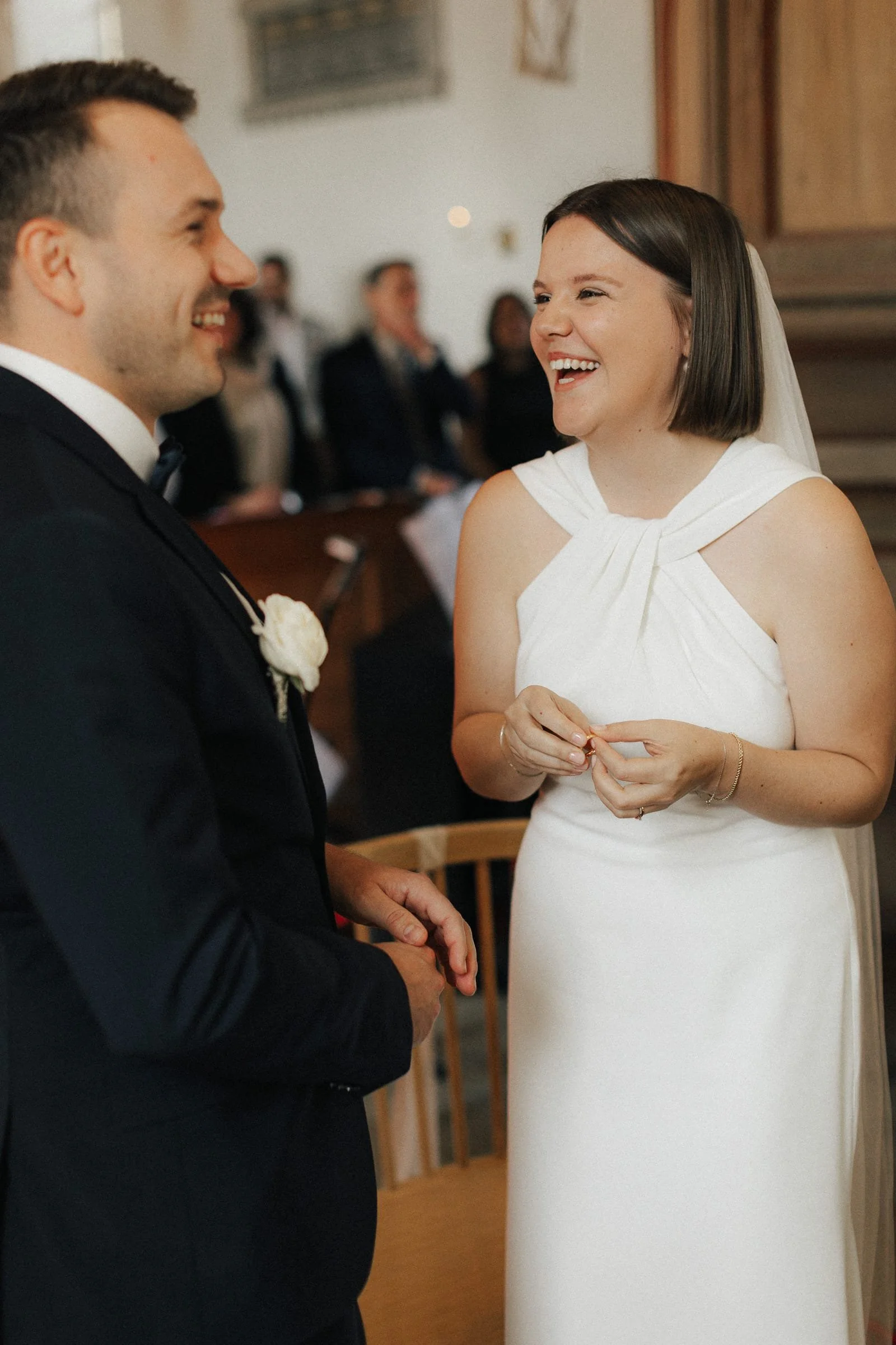 Ein Brautpaar bei einer Hochzeit, das sich liebevoll ansieht und lacht. Der Mann trägt einen schwarzen Anzug mit Fliege, die Frau ein weißes Kleid mit Schulterschleifen.