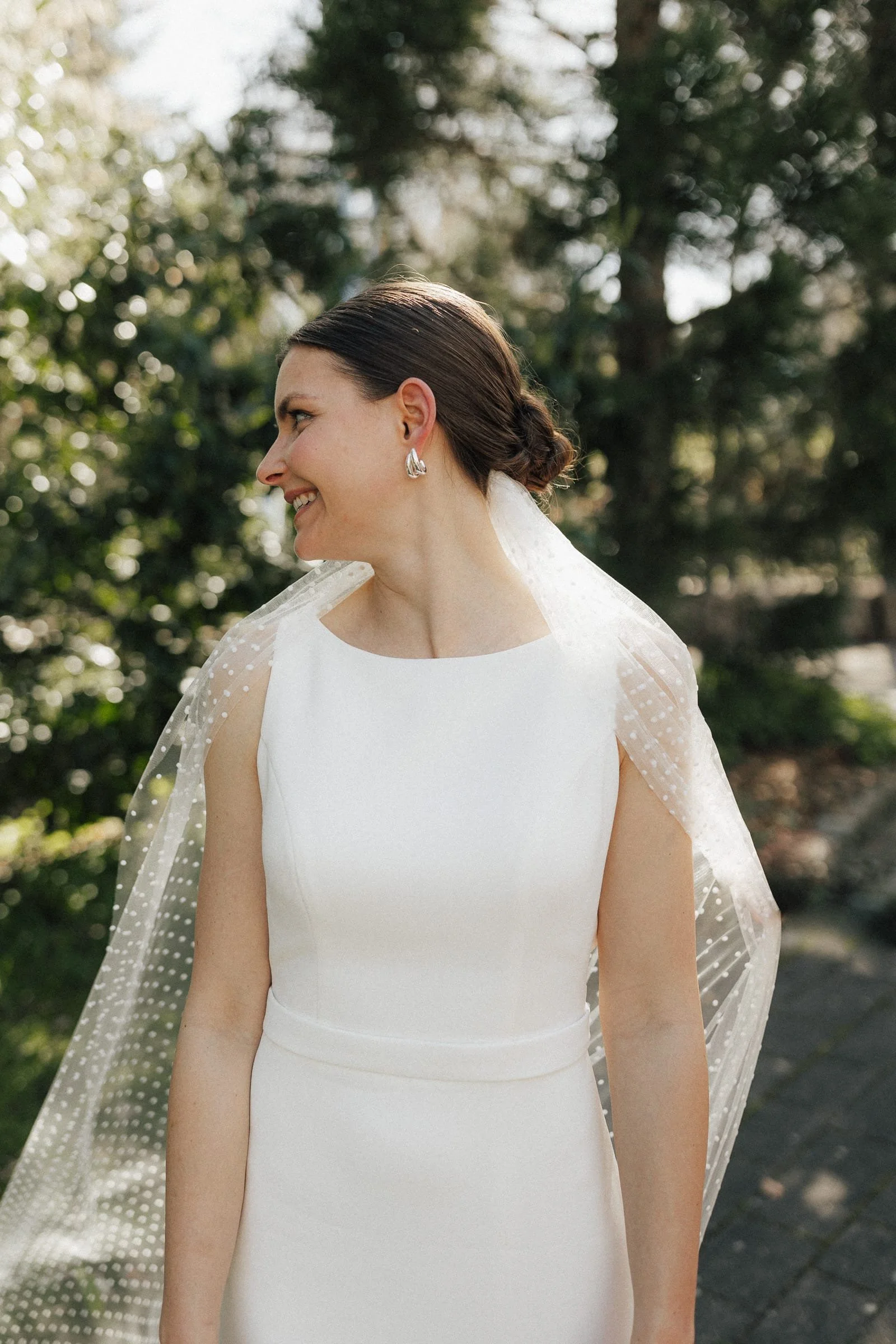 Eine smiling Braut in einem weißen Kleid mit einer transparenten Schleife mit Punkten. Sie trägt silberne Ohrringe und hat ihre Haare zu einem Dutt frisiert, während sie nach links schaut, im Freien mit Bäumen im Hintergrund.