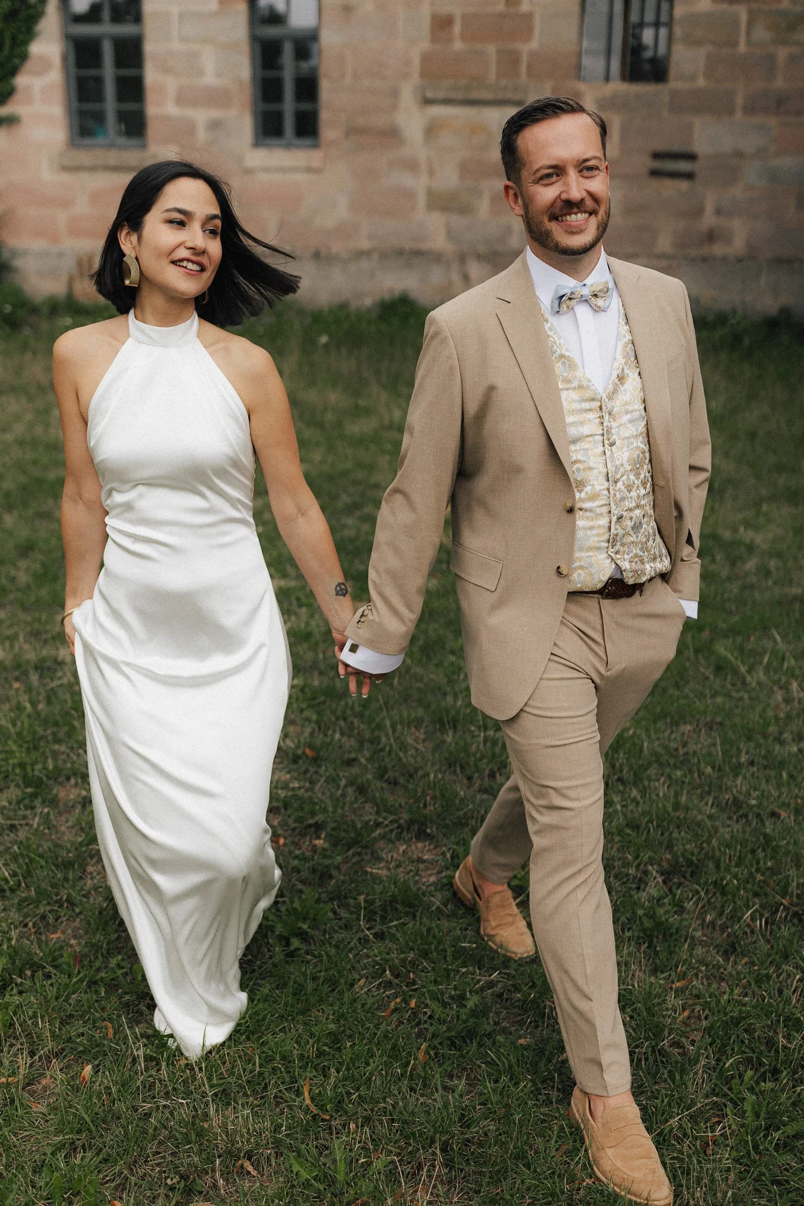 Ein lächelndes Paar im Hochzeitsoutfit spaziert auf einer Wiese vor einem alten Backsteingebäude.