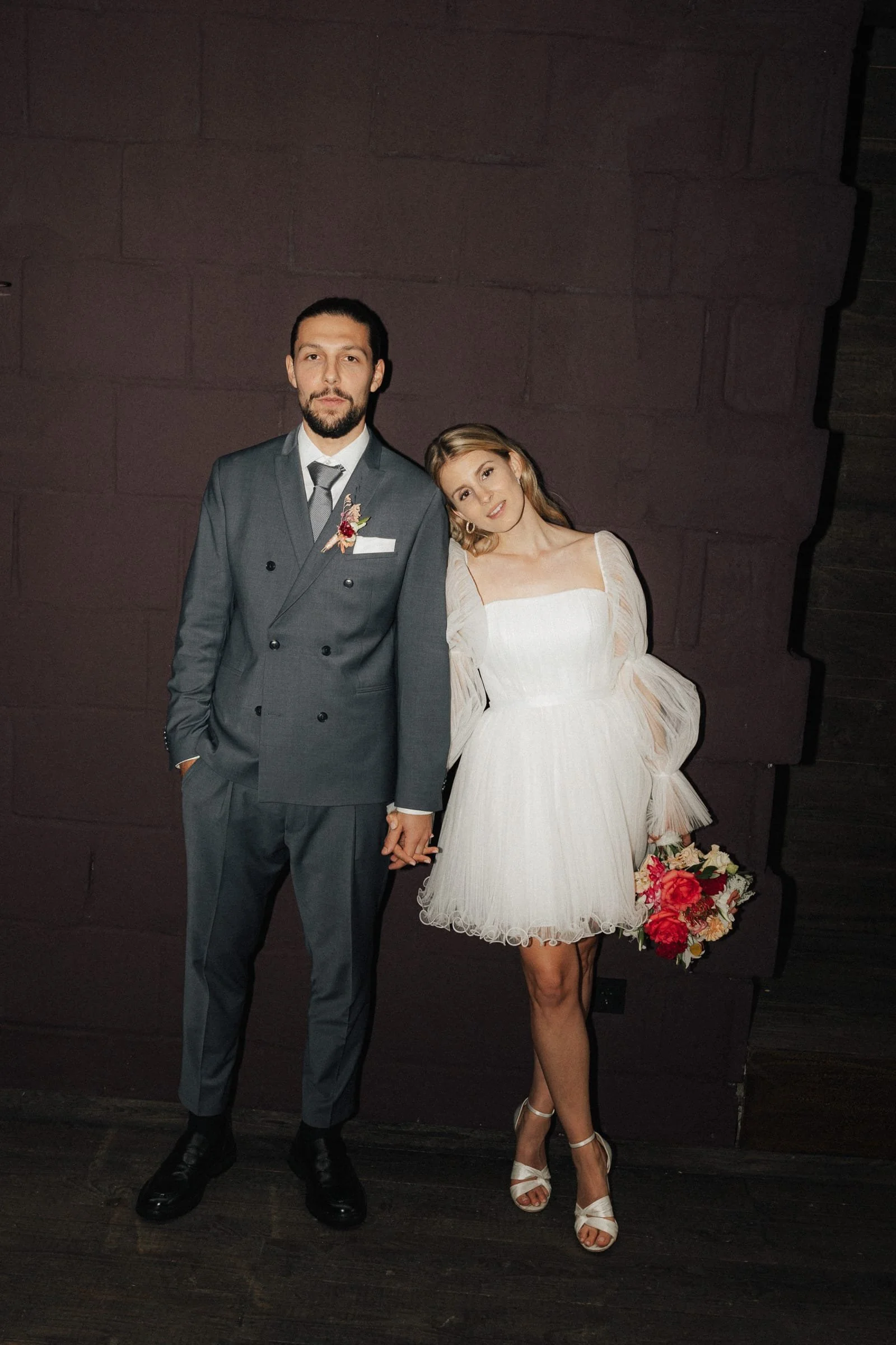 Ein Mann in einem grauen Anzug und eine Frau in einem weißen Kleid halten sich an den Händen und posieren vor einer dunkelroten Wand.