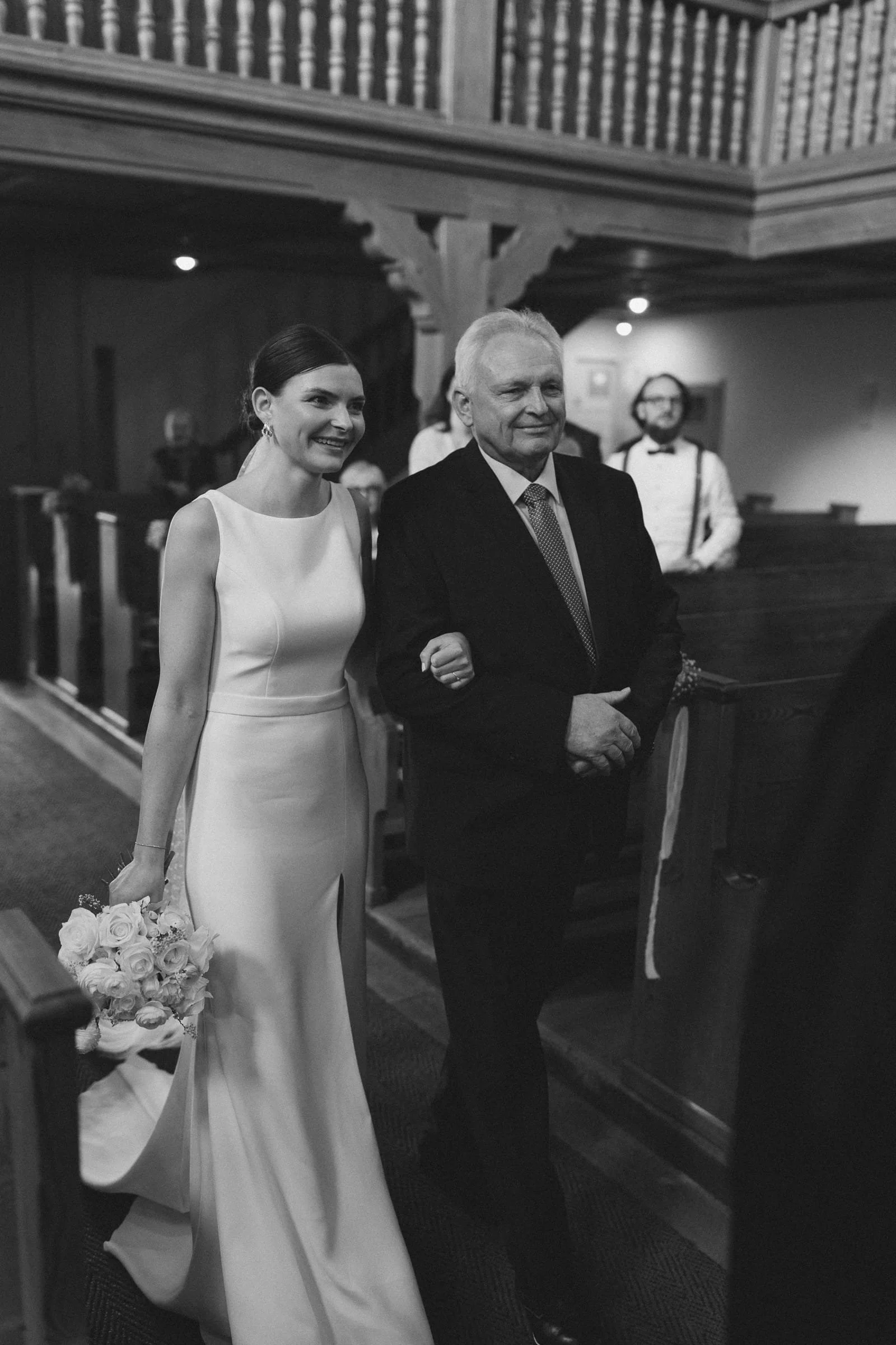 Braut in weißem Kleid läuft mit einem älteren Mann in Anzug in einer Kirche, hält einen Blumenstrauß in der Hand.