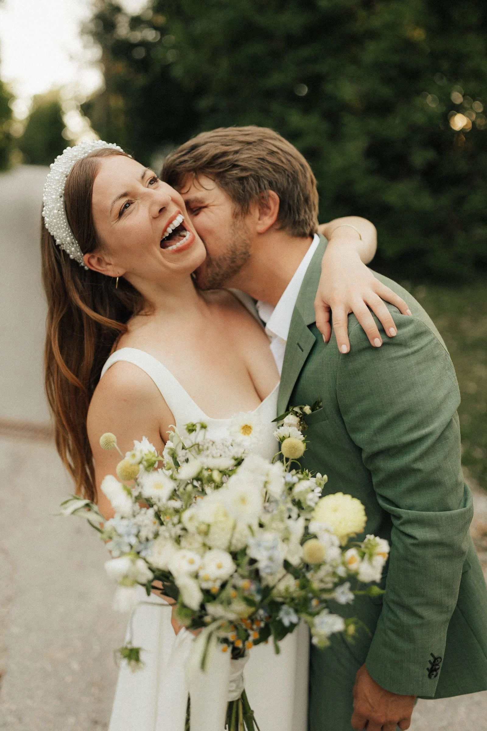 Ein glückliches Brautpaar umarmt sich bei der Hochzeit, die Braut trägt ein weißes Kleid und einen Perlenhaarreif, der Bräutigam trägt einen grünen Anzug, im Hintergrund grüne Bäume.