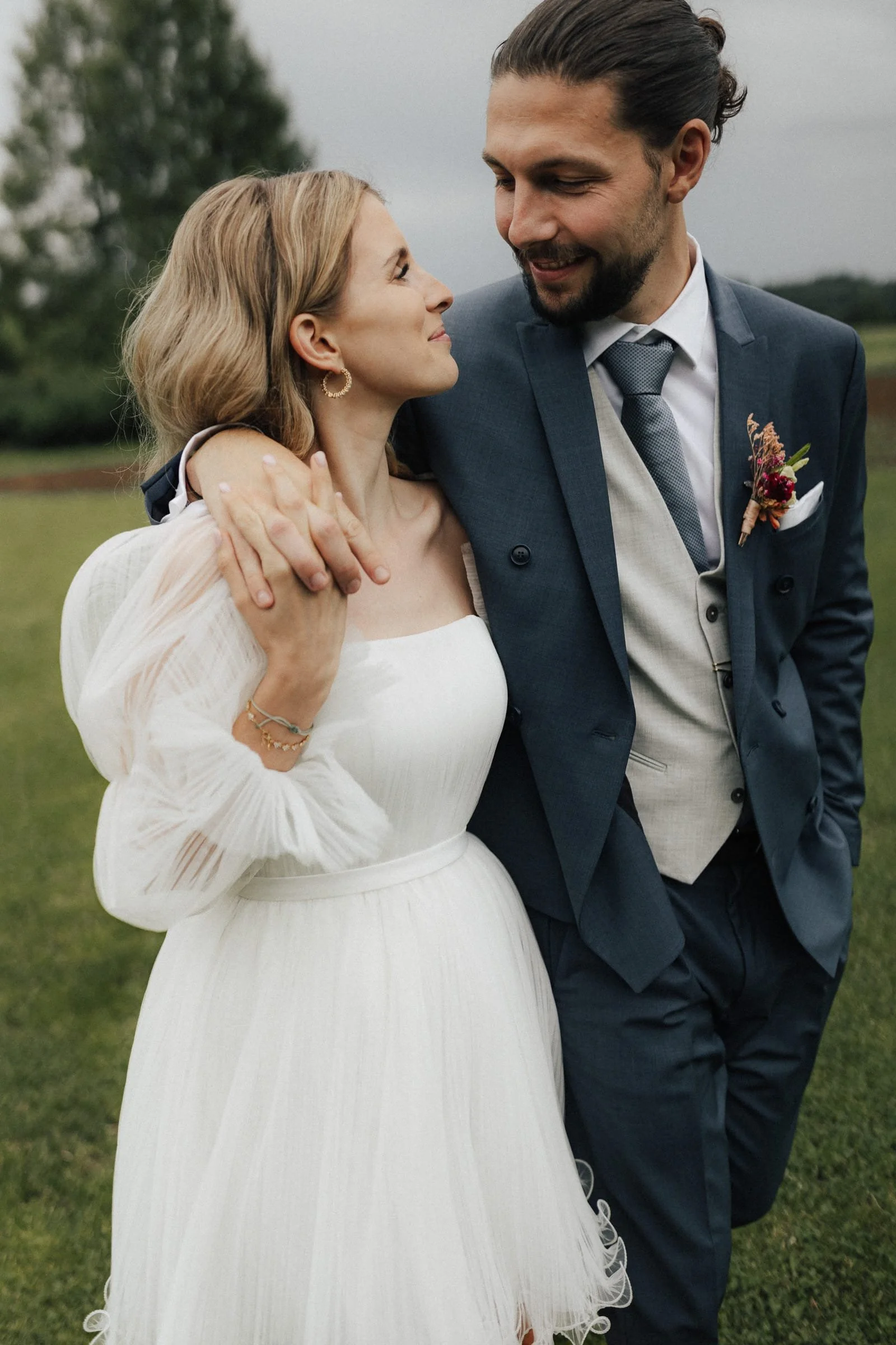 Ein frisch verheiratetes Paar, das sich liebevoll anschaut, im Freien bei bewölktem Himmel, mit einem grünen Feld im Hintergrund.
