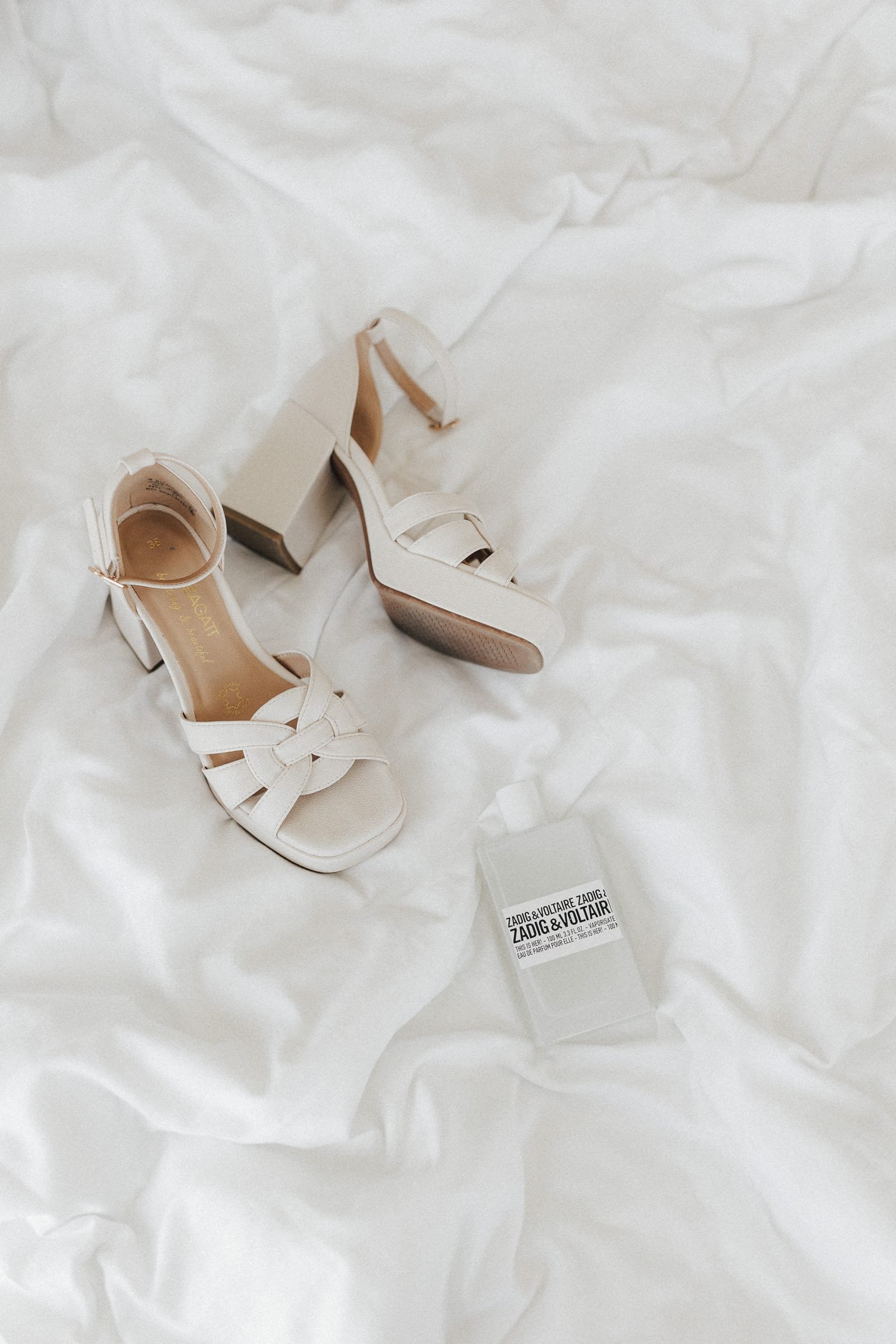 Weiße hohe Sandaletten mit Blockabsatz und Zierknöchelriemchen, ein Parfümfläschchen "Zadig & Voltaire" und weiße Seidenpapieraufnahmen.