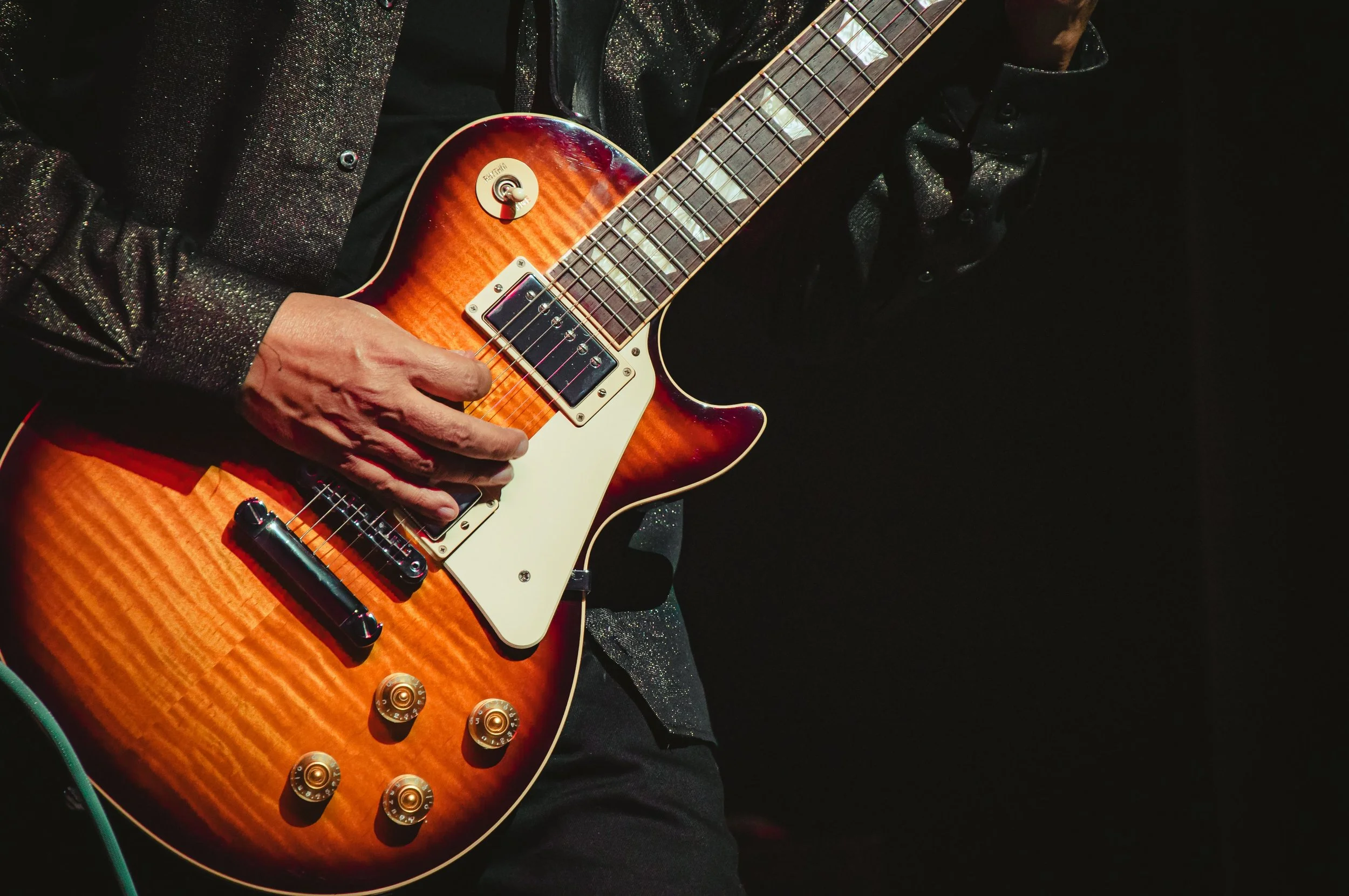 Close up of an electric guitar being played by a man in a leather jacket and black troysers 