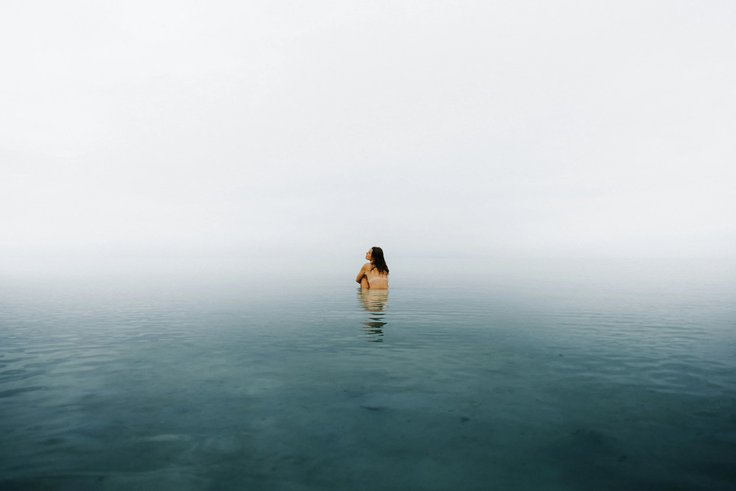 A person with long hair appears to be topless, standing in calm water with a foggy or cloudy sky in the background.