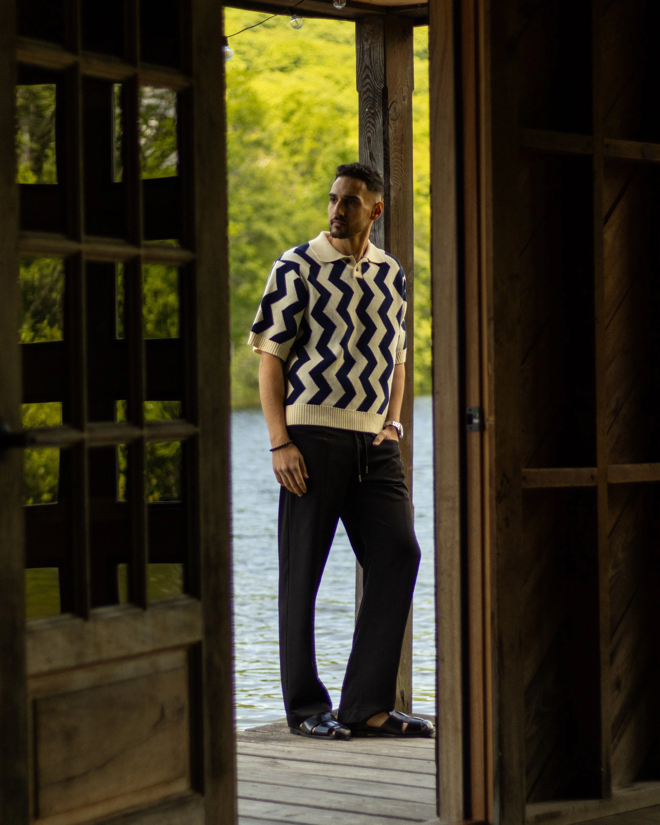 A man with dark hair, a beard, and light skin stands on a wooden dock by a body of water, wearing a zigzag patterned knit sweater, black pants, and sandals, as seen through a small open door in a wooden structure surrounded by greenery.