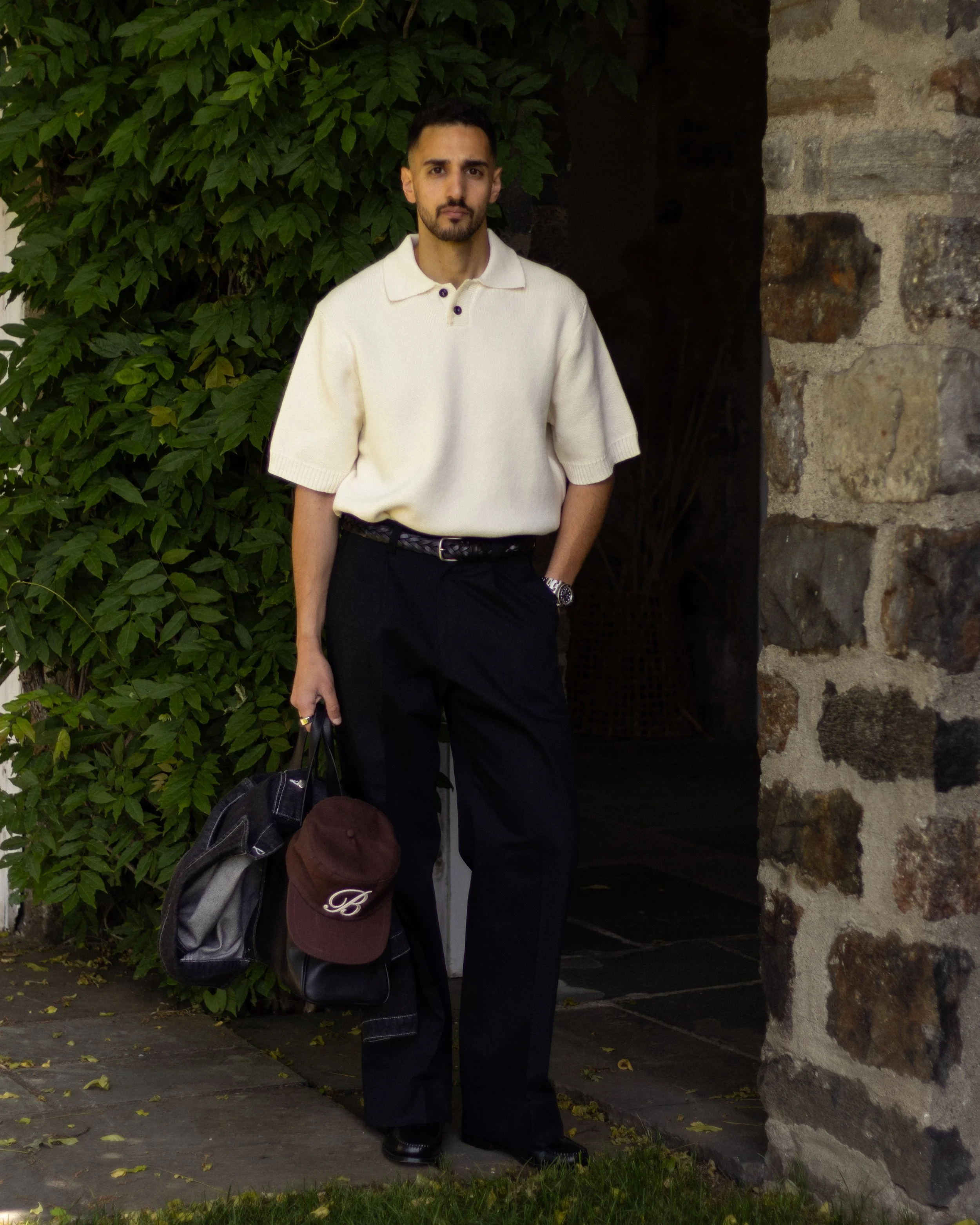 A man with dark hair and beard wearing a cream-colored short-sleeve polo shirt and black pants, standing next to a stone wall and green leafy bushes.