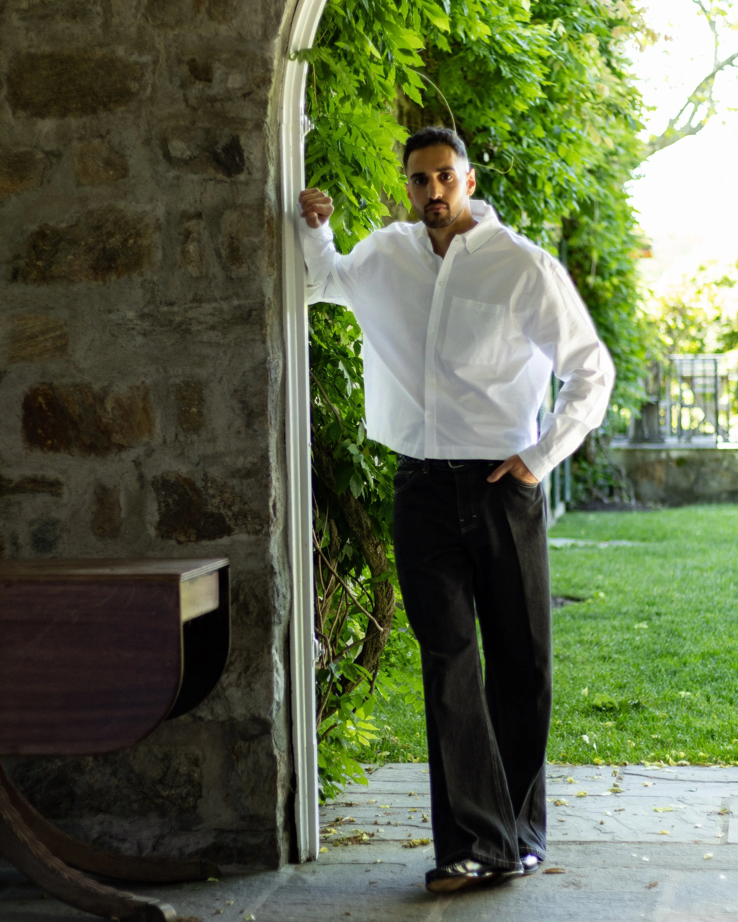 A man in a white dress shirt and black pants stands outside near a stone wall, holding onto a white door frame with greenery and trees in the background.