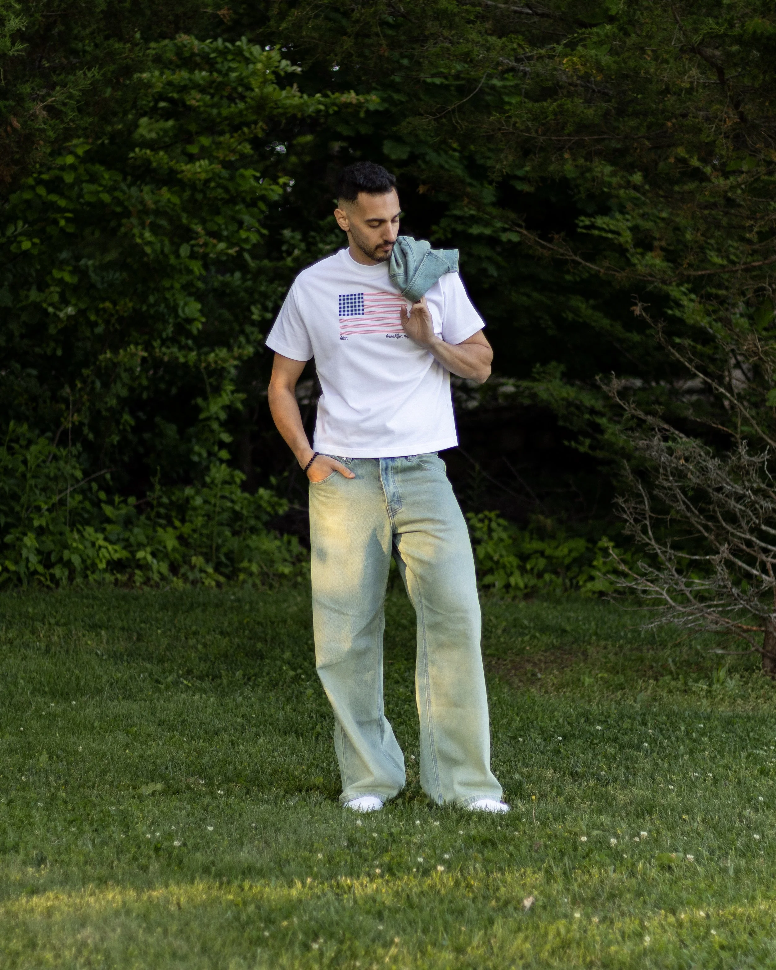 A man standing on grass in front of trees, holding a gray shirt over his shoulder, wearing a white t-shirt with a graphic of the American flag and light-colored jeans.