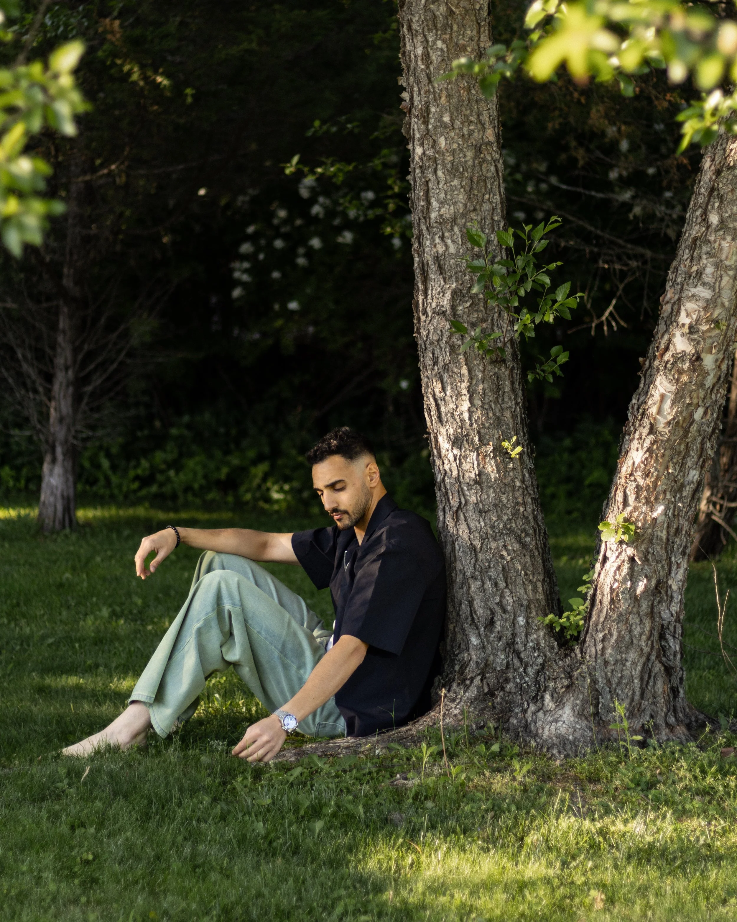 A man sits under a large tree in a grassy area, with his eyes closed, relaxed with one arm resting on his knee. He's wearing light-colored pants, a dark shirt, a watch, and a bracelet, and is surrounded by greenery and other trees.