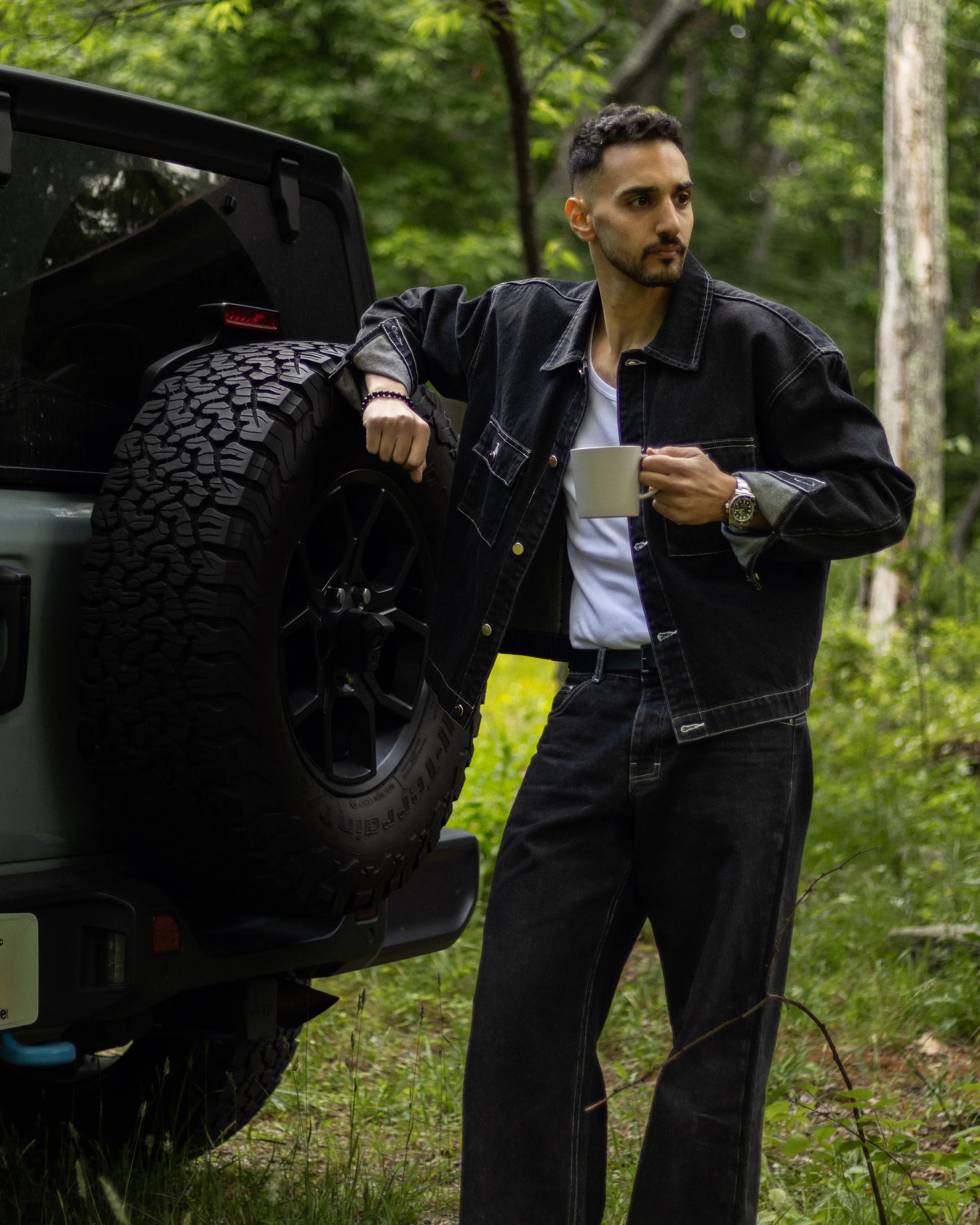 A man standing outdoors in a wooded area, holding a mug, leaning against a vehicle with a spare tire on the back.