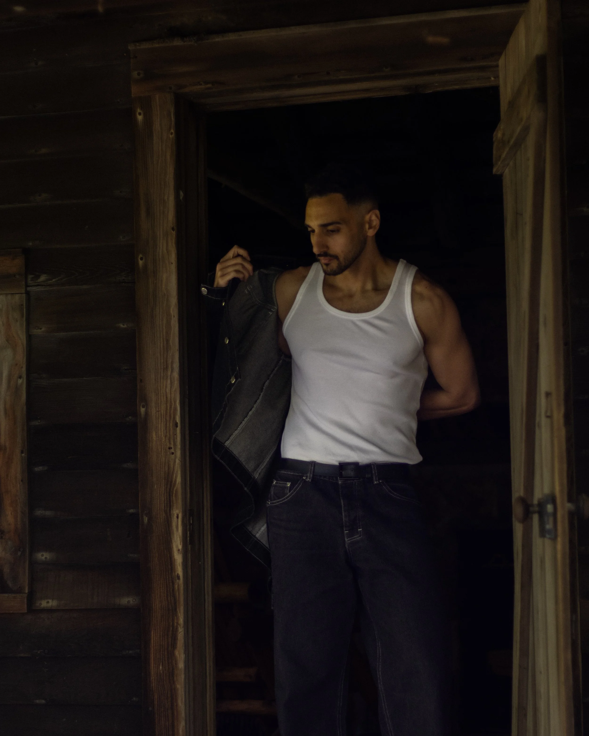 A man standing in a dark wooden structure, wearing a white tank top and jeans, holding a jacket over his shoulder and looking down.