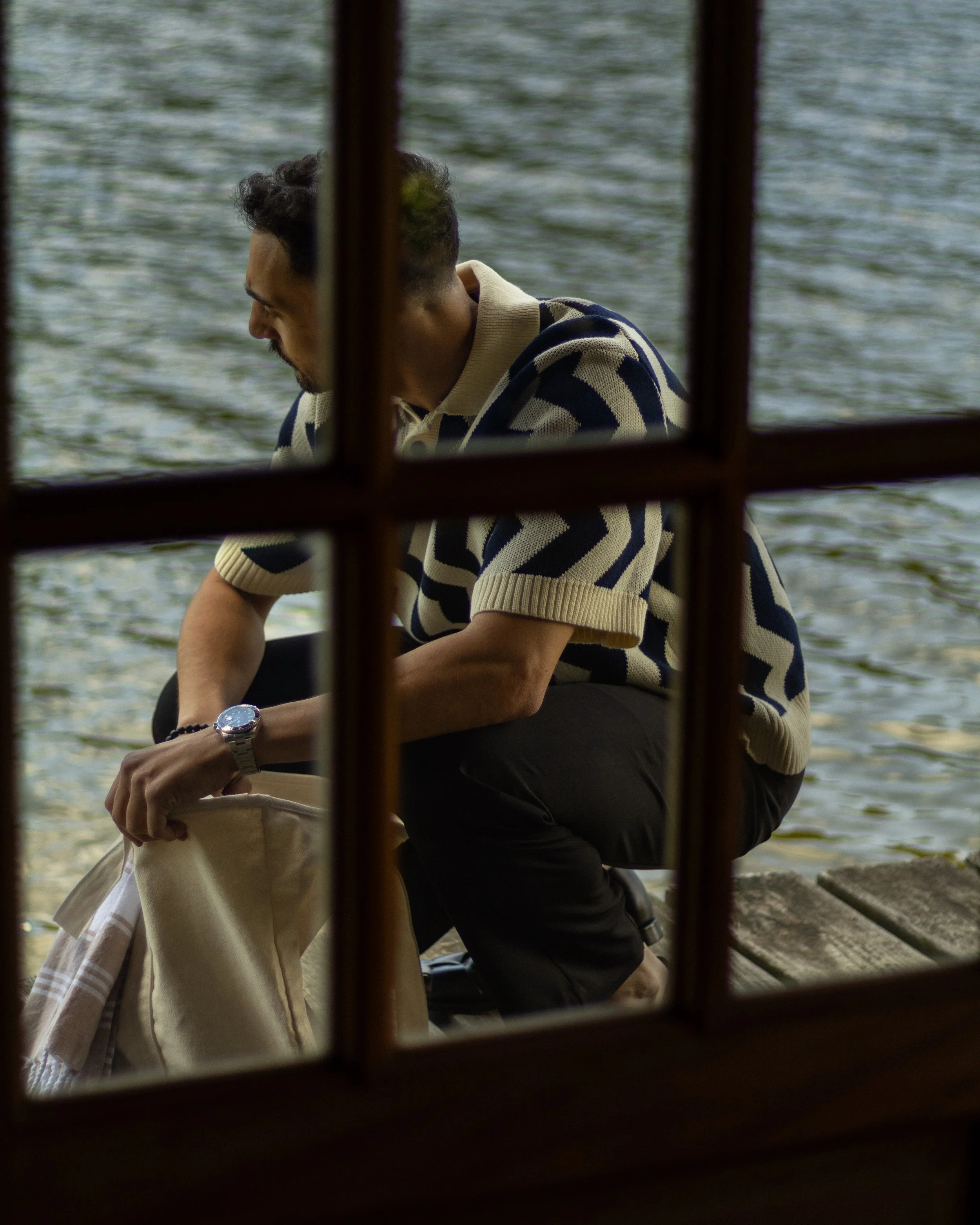 A man with dark, curly hair, a beard, wearing a striped short-sleeve sweater, is sitting outdoors by a body of water, seen through a window with wooden frame bars.