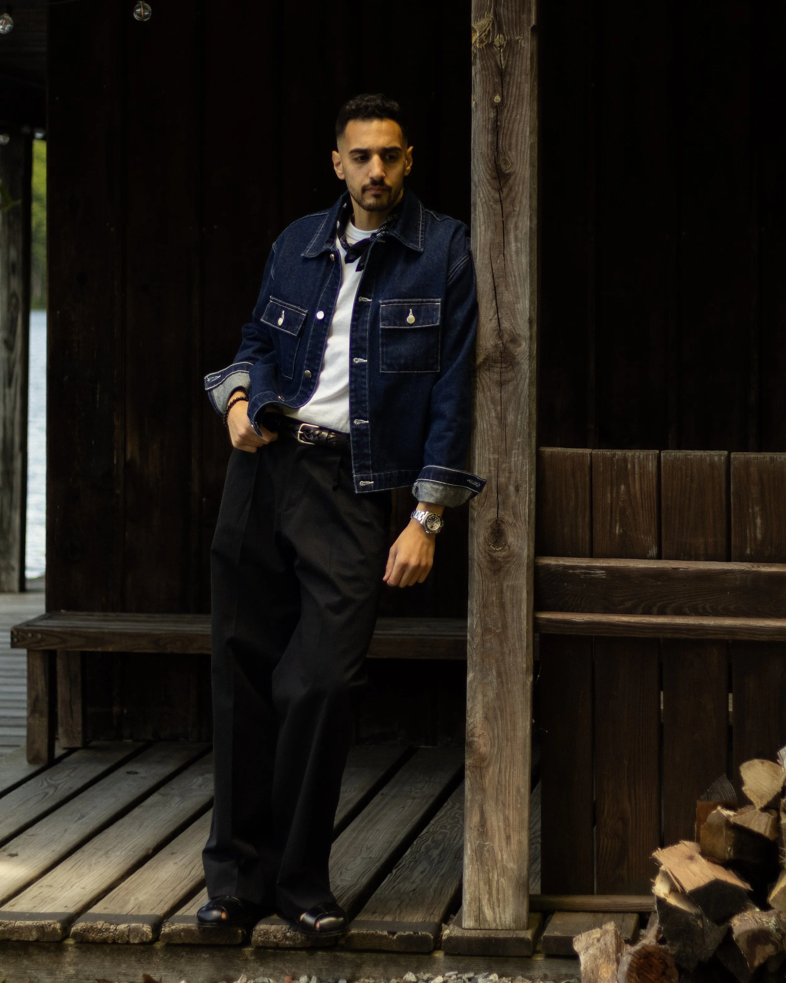 A young man in a denim jacket, white shirt, and black pants stands on a wooden dock next to a dark wooden wall, with trees in the background. He is looking down, with his hands in his pockets, wearing a watch and bracelets.