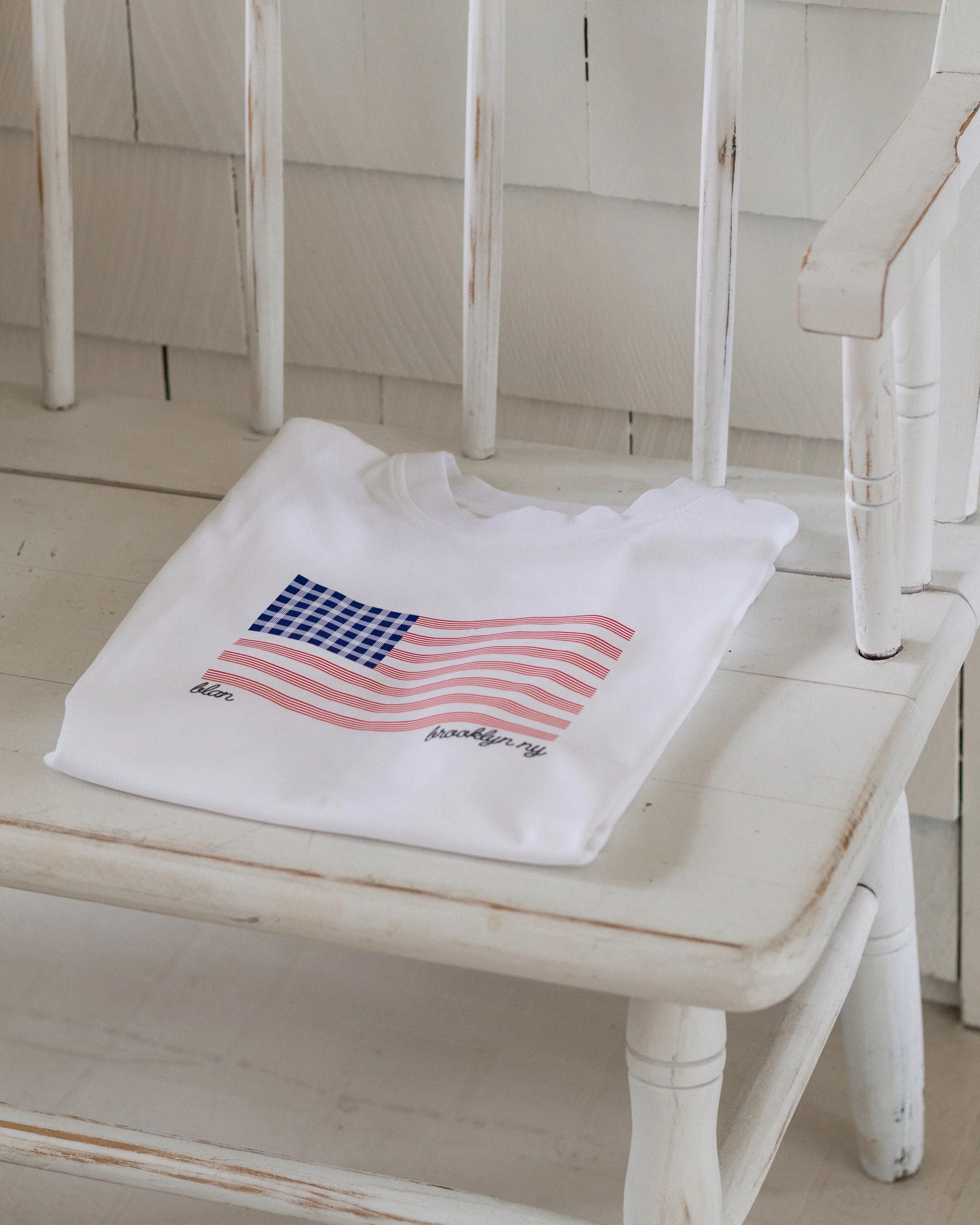 A white t-shirt with a patriotic American flag design, including the words 'bless' and 'brooklyn ny', lying on a white wooden surface.